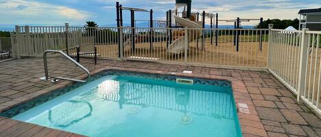 A swimming pool with a slide, surrounded by a fenced area and a view of the sea.