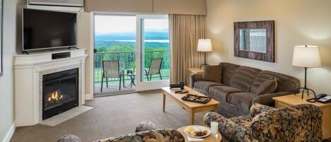 A hotel room with a fireplace, a television, a sofa, a coffee table, and a balcony with a view of greenery.