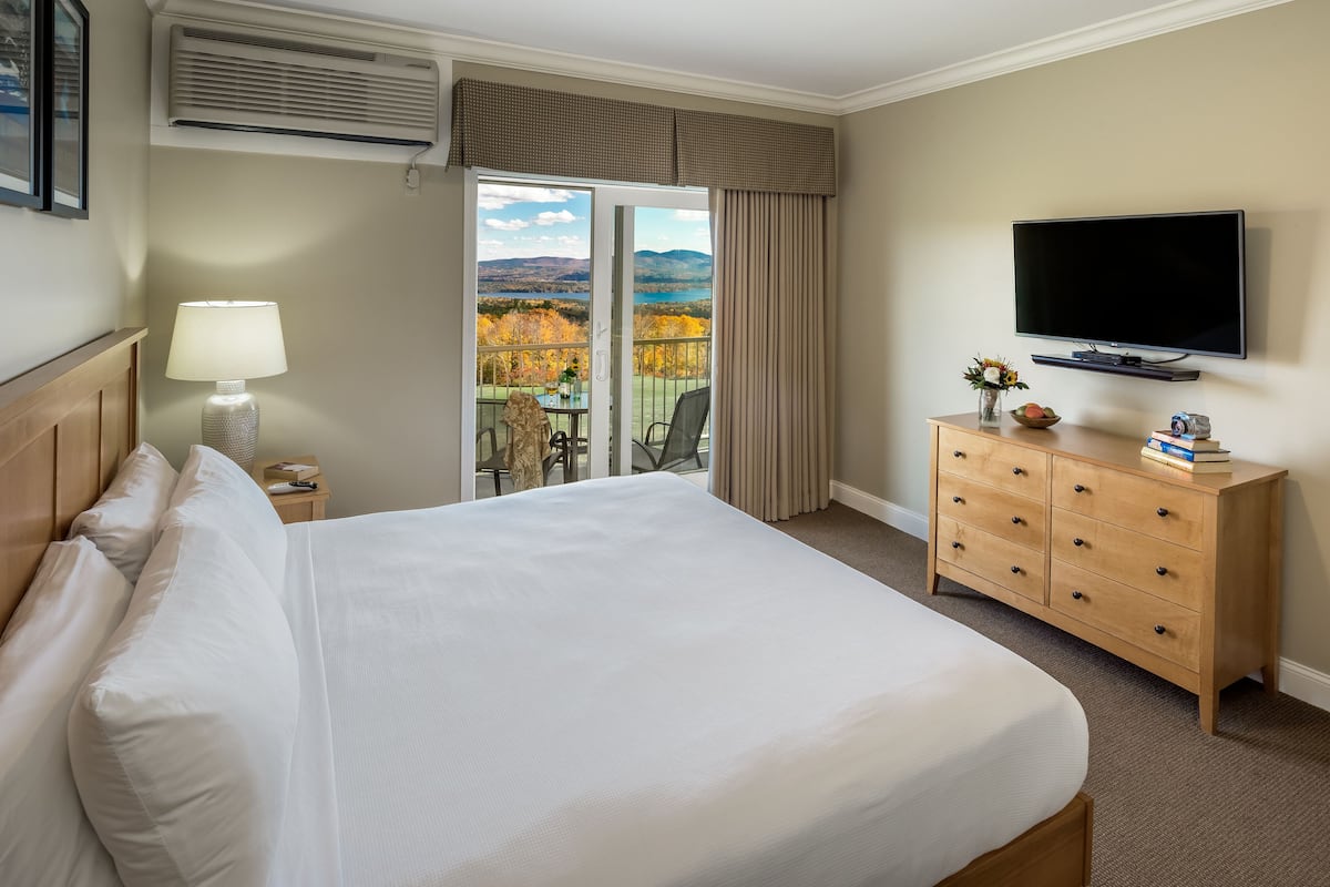 A hotel room with a large bed, a TV, a dresser, and a balcony with a scenic view.