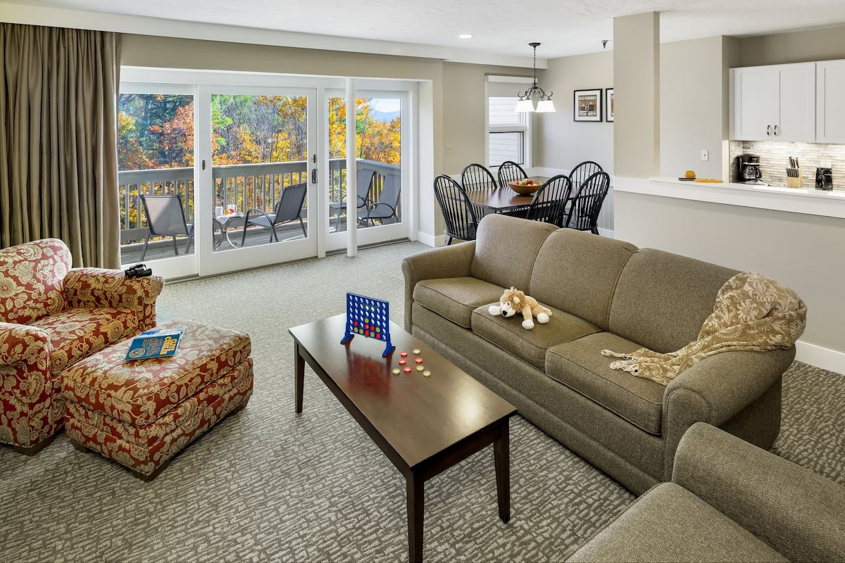 A living room with a sofa, chairs, a coffee table, and a view of autumn trees through large sliding glass doors.