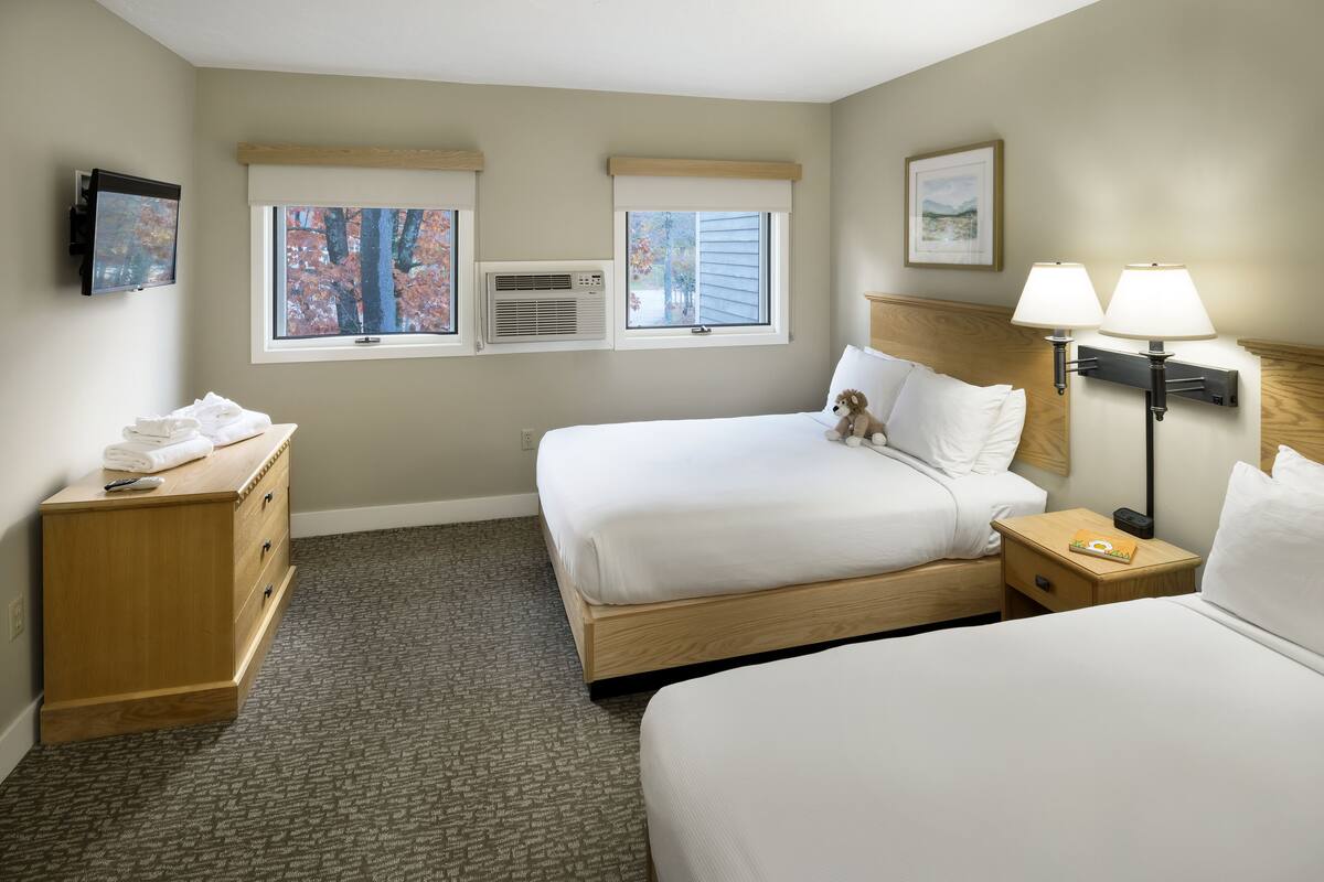 A hotel room with two beds, a wooden dresser, a TV, and a window with a view of trees.