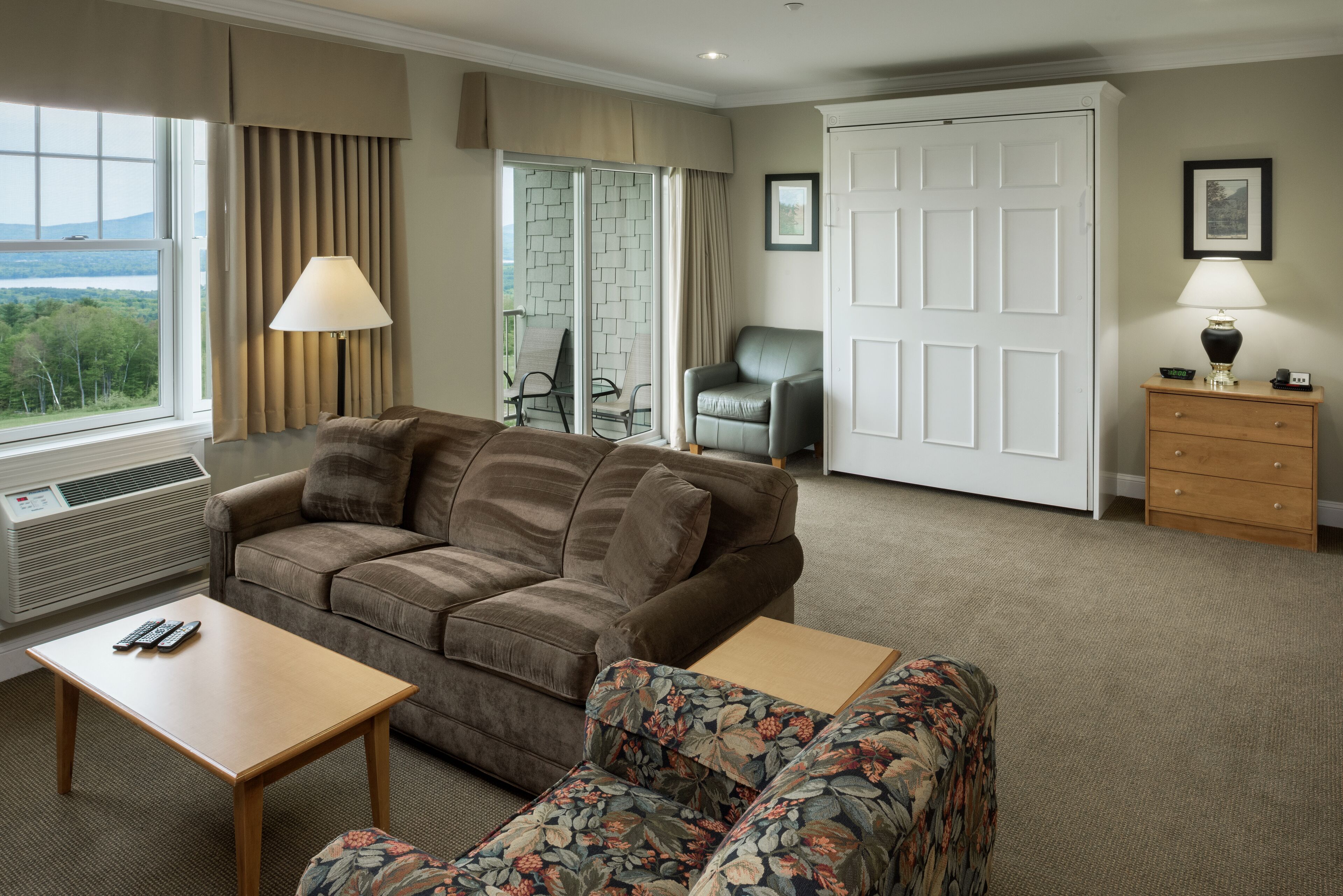 A hotel room with a sofa, a floral chair, a wooden table, a lamp, a wardrobe, and a view of the outdoors.