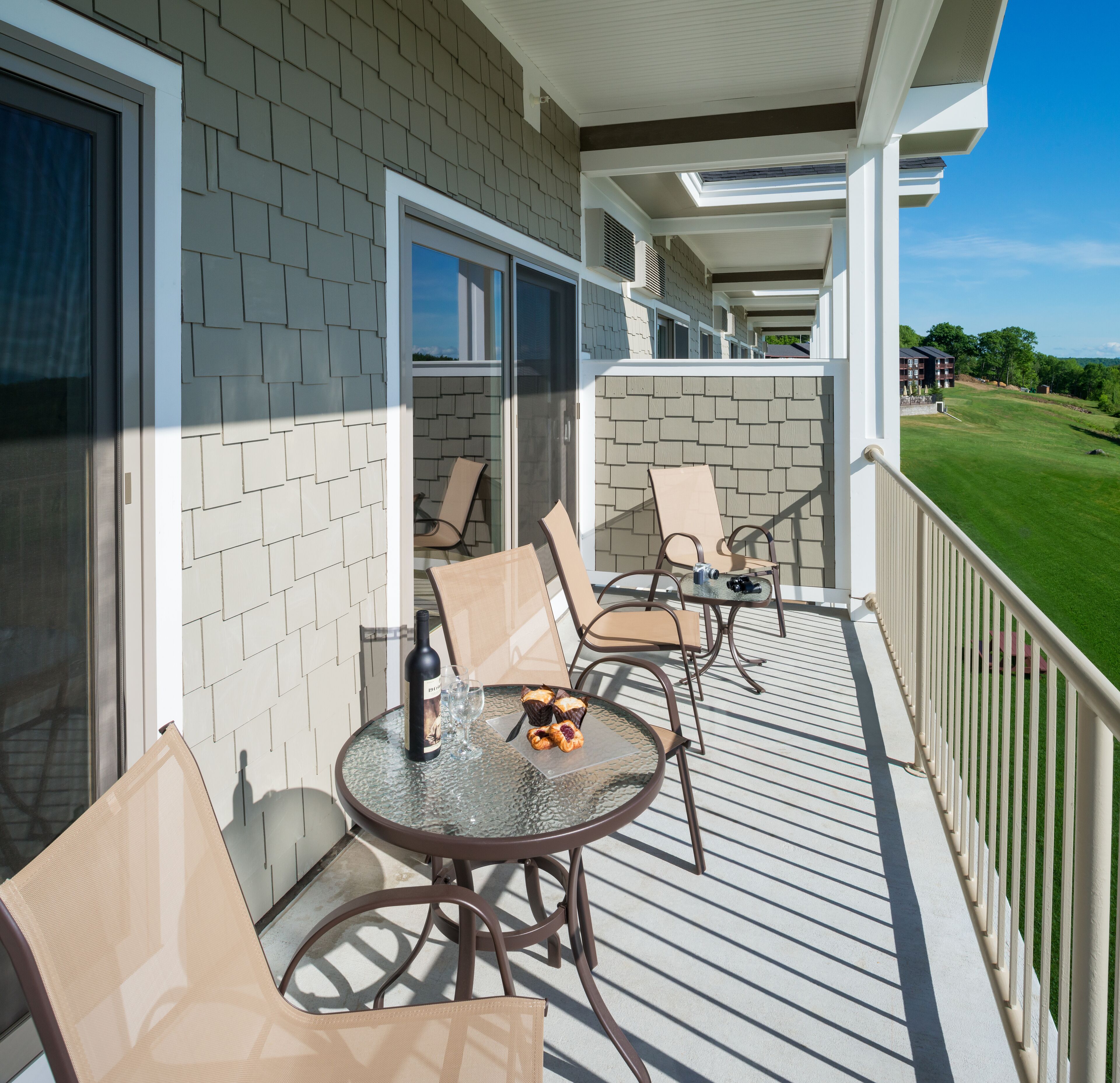 A balcony with outdoor seating, a table with a bottle and glasses, and a view of a green landscape.