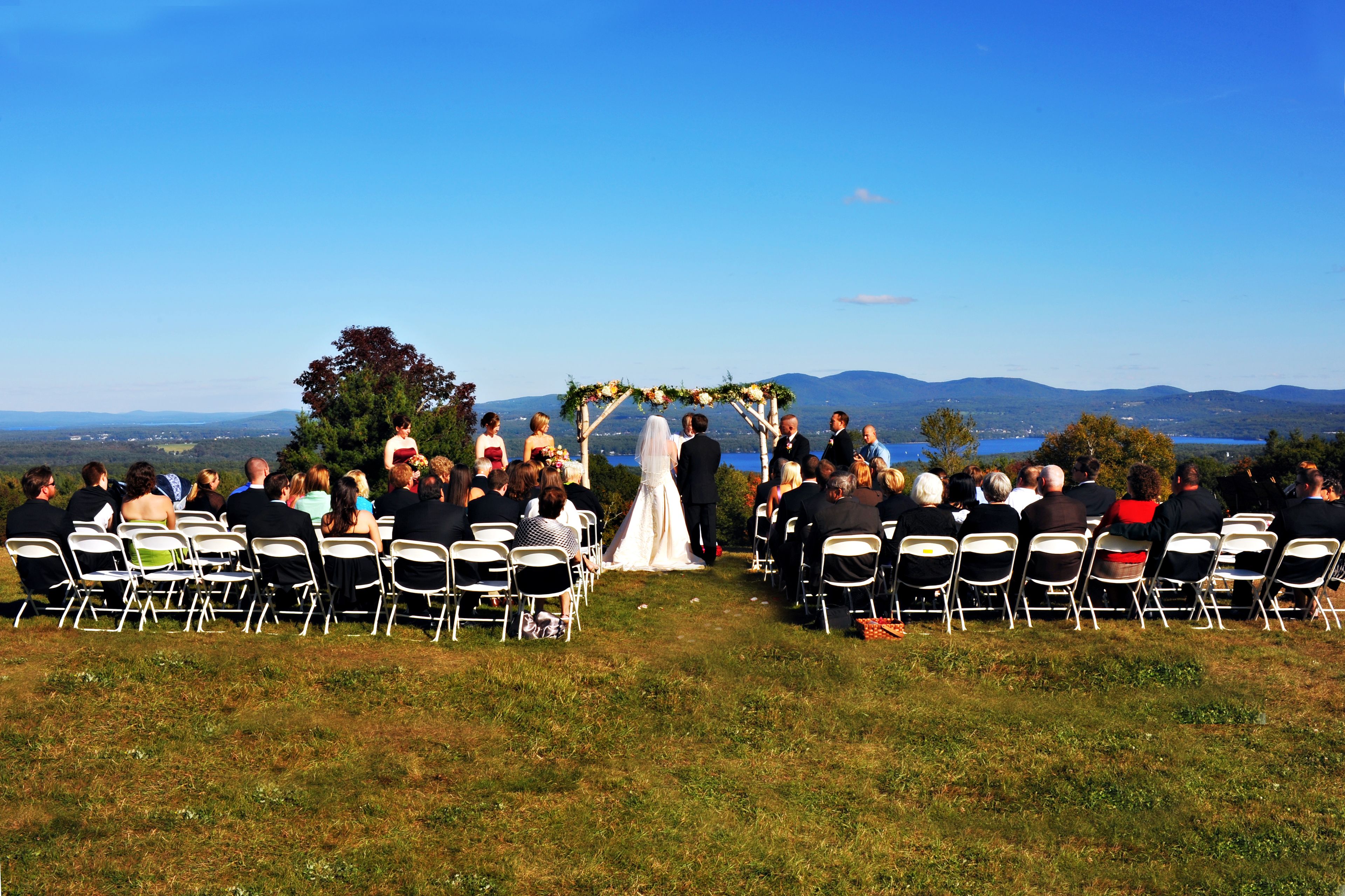 A wedding ceremony outdoors with guests seated on white chairs facing the aisle.