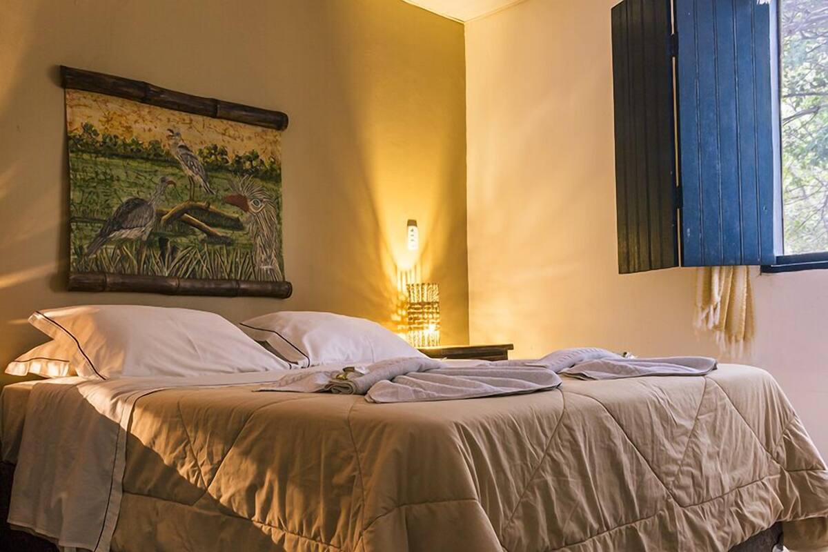 Una cama con colcha color crema, un cuadro de pájaros colgado en la pared, un aplique de pared y una ventana con persianas oscuras.
