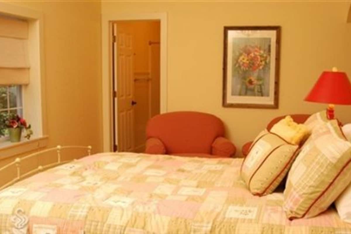 A bedroom with a bed, a red chair, a painting on the wall, and a window with flowers.
