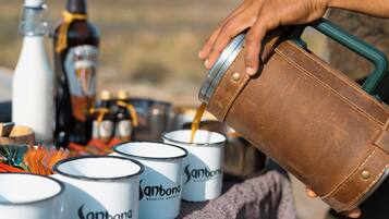 A person pouring a liquid from a brown leather container into white enamel mugs labeled "Sanbona Wildlife Reserve."