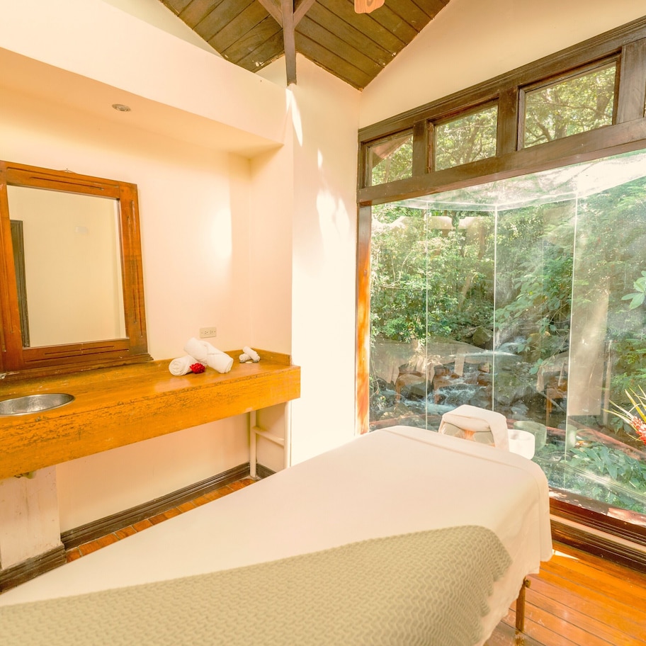 A room with a wooden vanity, a mirror, a sink, a bed, and a large window with a view of greenery.