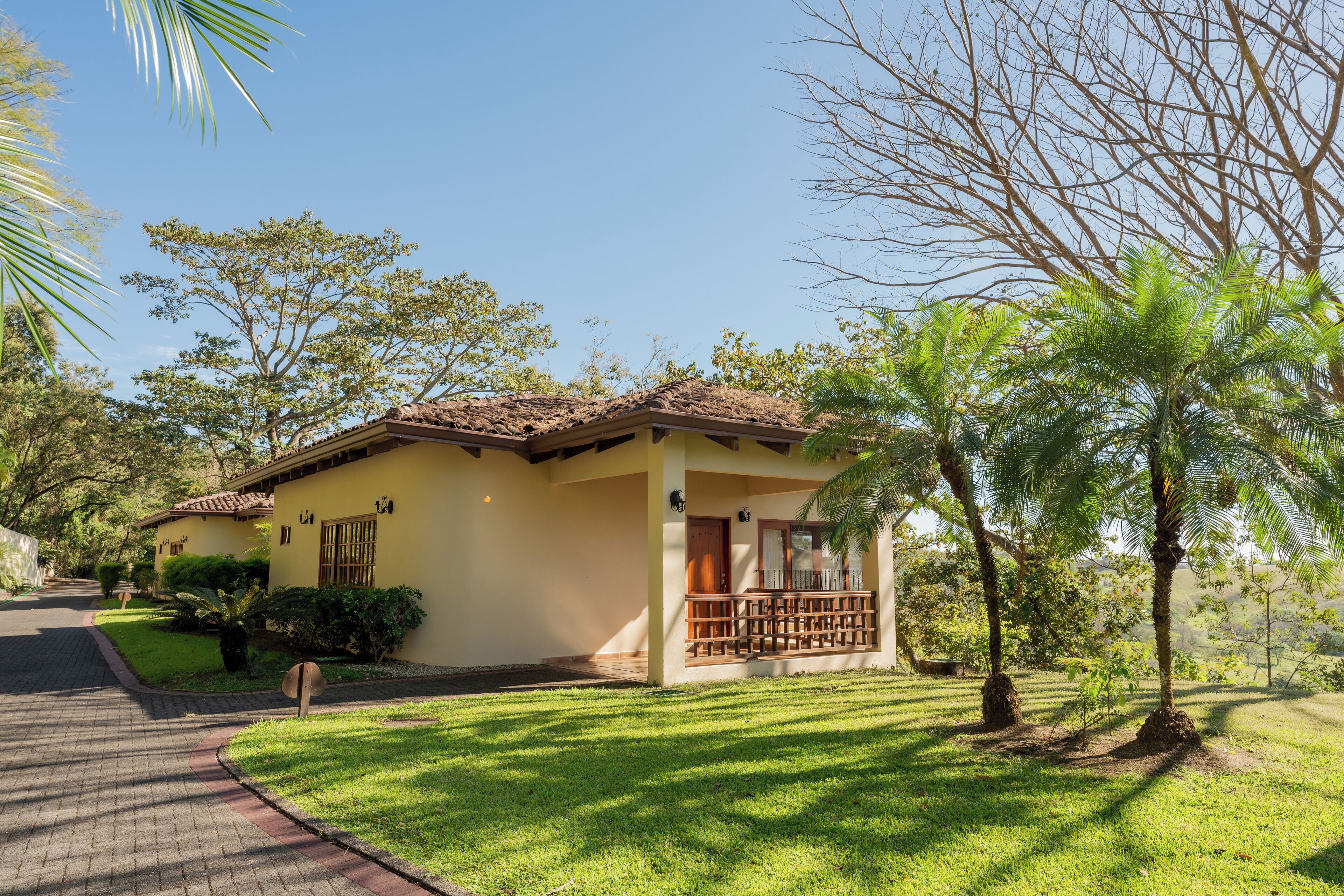 Um edifício amarelo com telhado de azulejos, cercado por vegetação e um caminho pavimentado.
