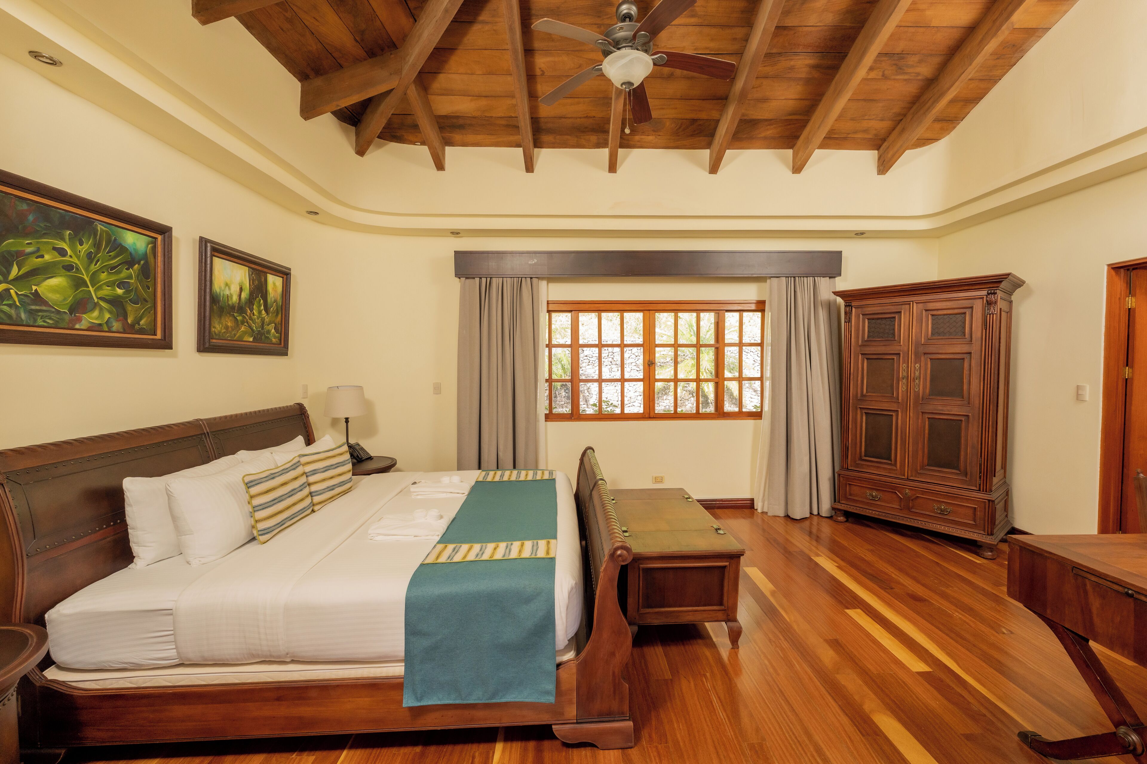 Un dormitorio con techo de madera, una cama con colcha verde, una mesita de noche, un banco de madera, un armario y una ventana con cortinas.