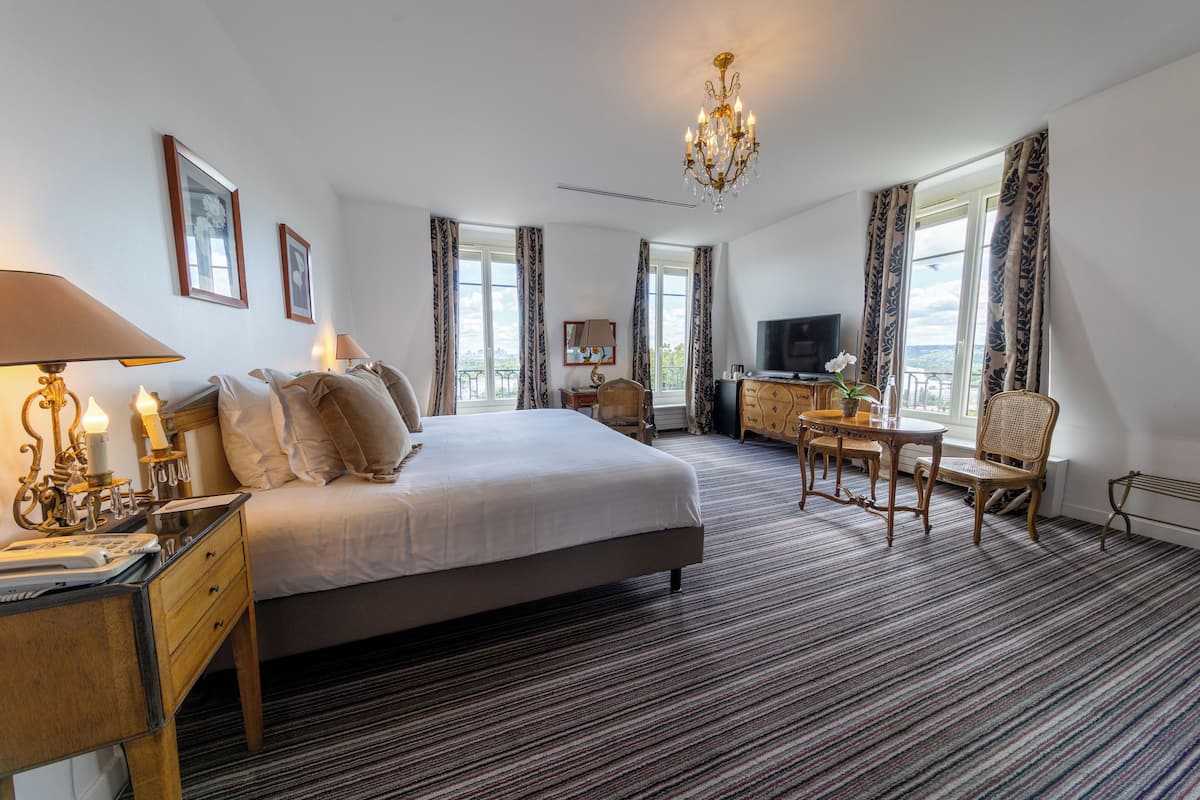 A hotel room with a large bed, a desk with a lamp, a television, and a view of the city through the windows.