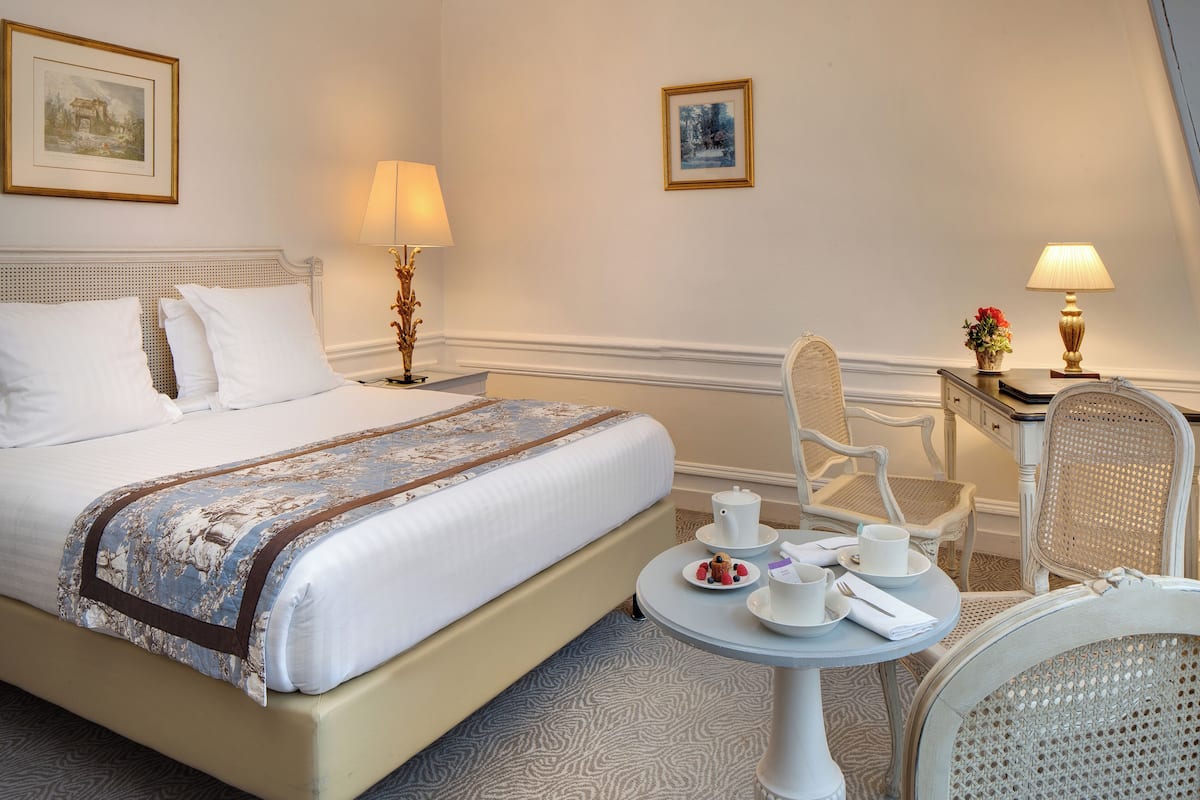 A neatly made bed with a floral bedspread, a bedside table with a lamp, a chair, and a small table with a teapot, cups, and a plate of strawberries.