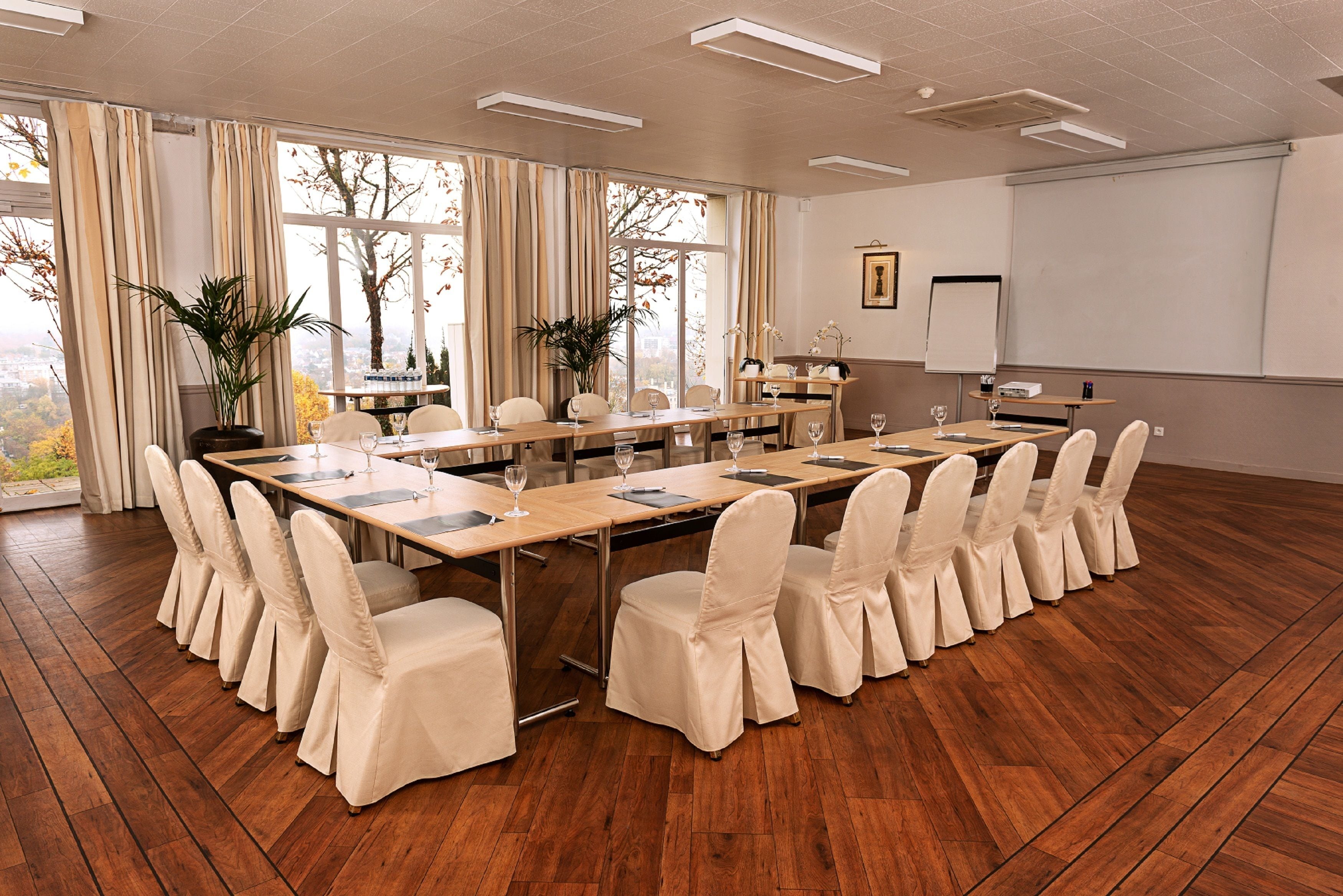 Une salle de conférence équipée d’une longue table, de chaises, d’un écran de projection et de grandes fenêtres avec des rideaux.