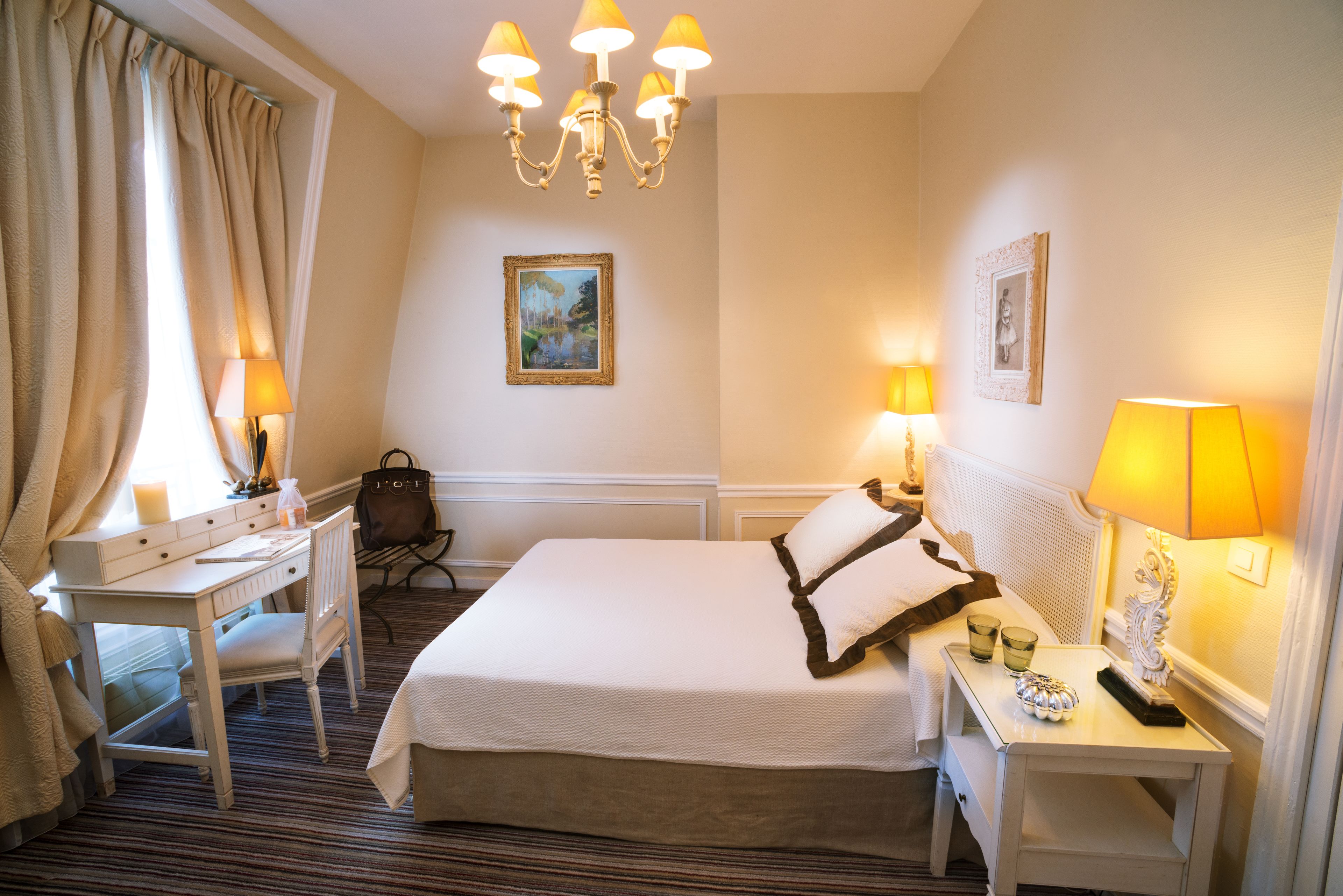 A hotel room with a bed, desk, chair, bedside tables, lamps, and a chandelier.