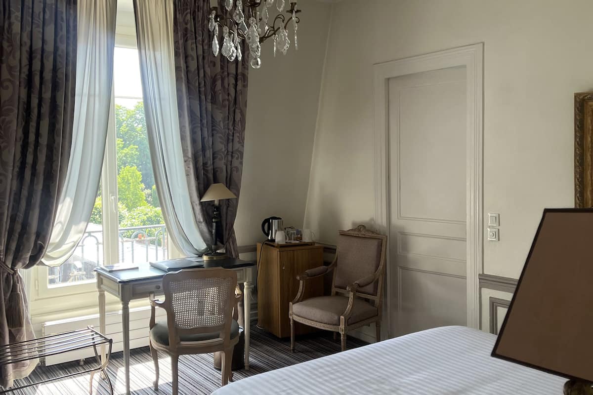 A hotel room with a large bed, a desk with a chair, a chandelier, and a view of greenery outside.