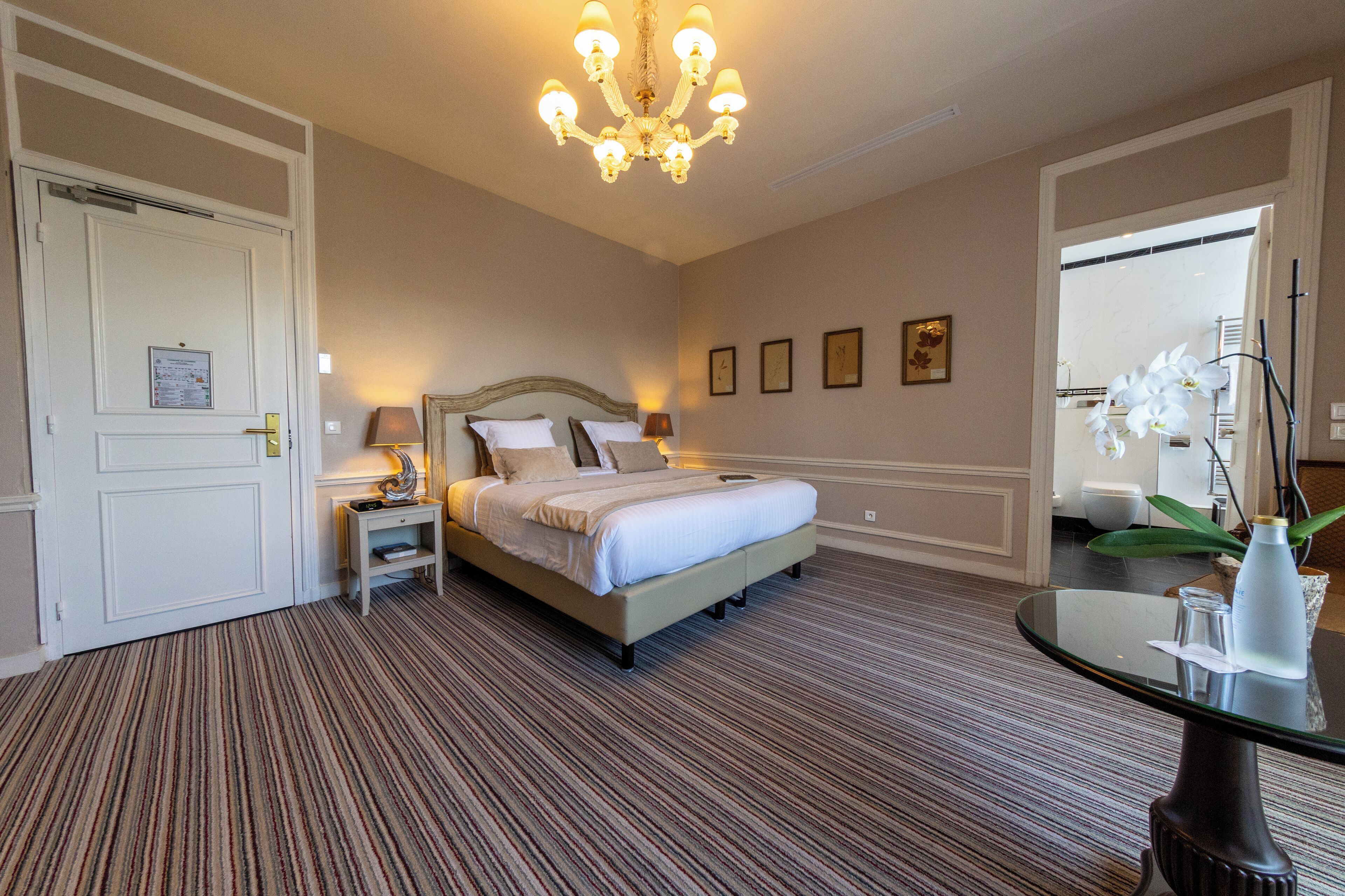 Habitación de hotel con una cama grande, una mesita de noche con lámpara, una araña y una pequeña mesa con un florero.