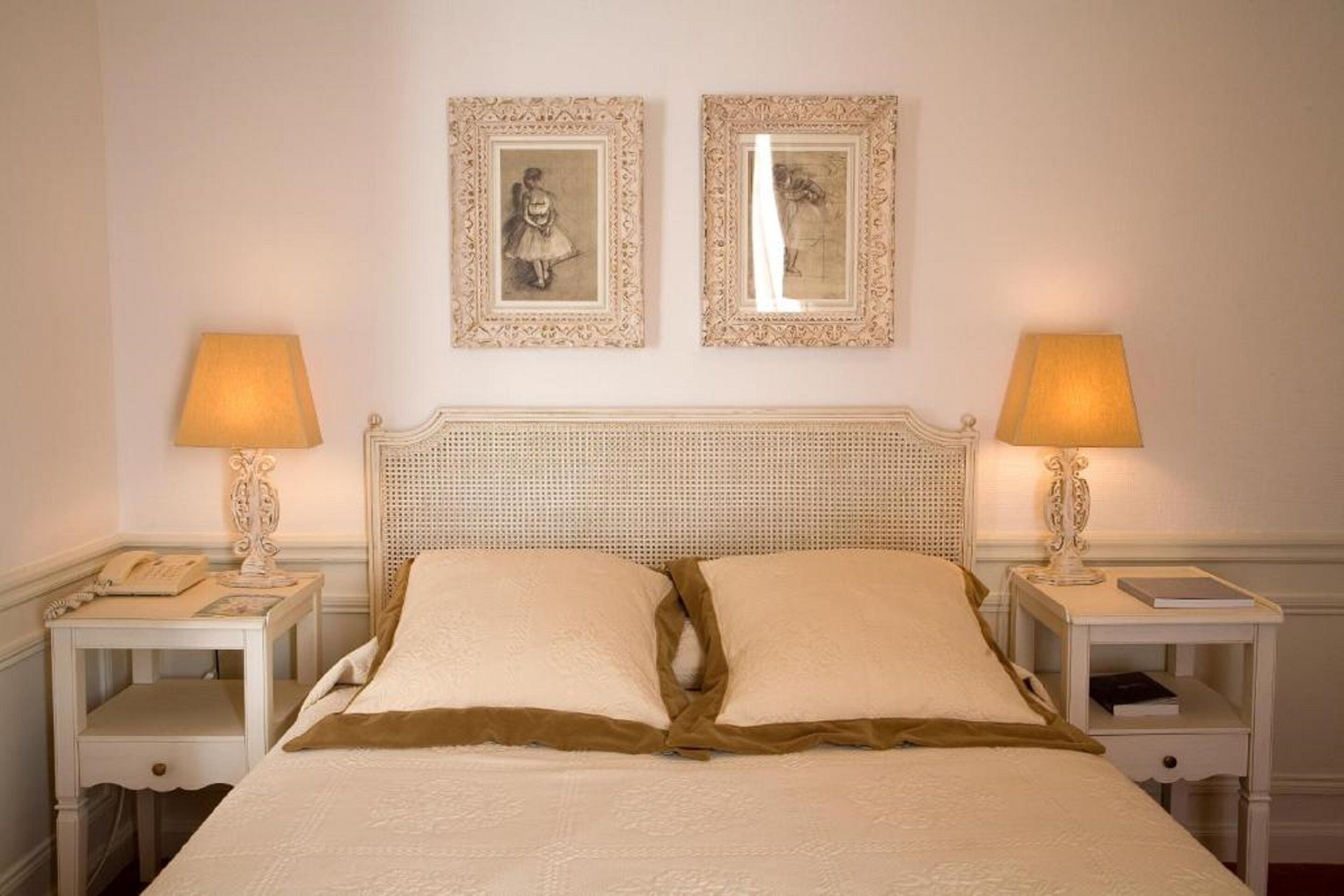 A neatly arranged bedroom with a classic headboard, two bedside tables with lamps, and two framed pictures on the wall.