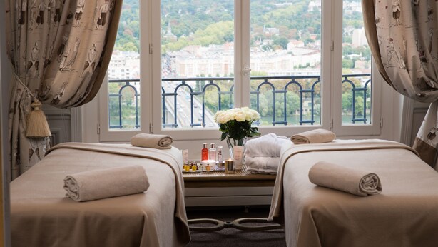 A hotel room with two beds, a window with a city view, a vase with flowers, and toiletries on the nightstand. - Pavillon Henri IV Hotel Restaurant Gastronomique Terrasse (Saint-Germain-en-Laye)