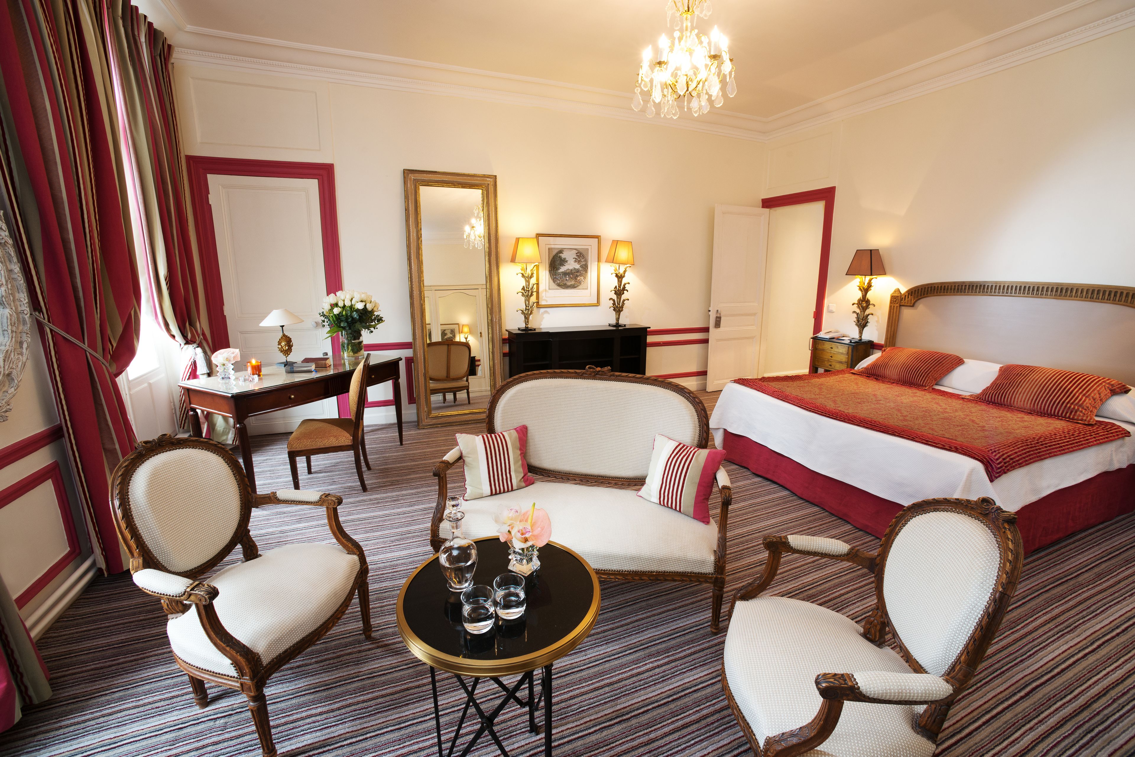 A hotel room with a large bed, two armchairs, a small table with a vase and glasses, a desk with a lamp, and a fireplace.