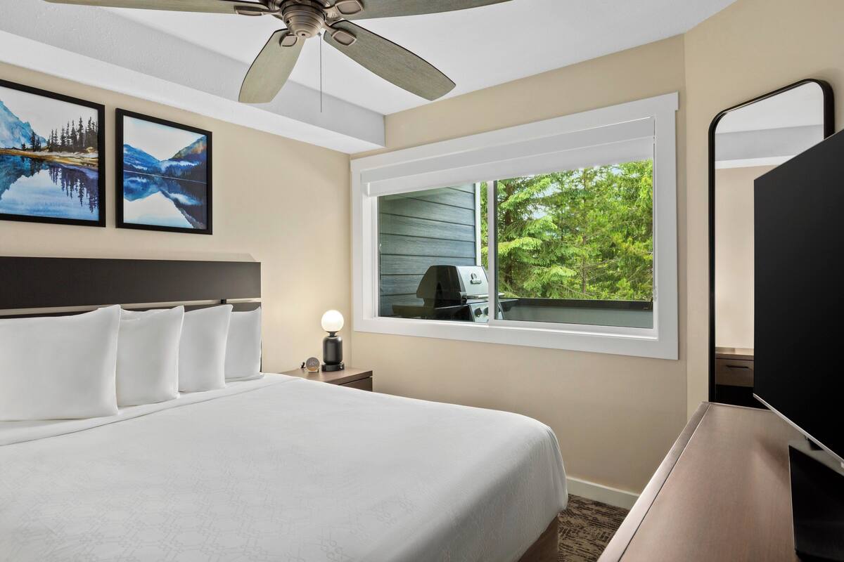 Ein Hotelzimmer mit einem Bett, einem Deckenventilator, einem Fenster mit Blick auf Grünflächen und zwei gerahmten Bildern an der Wand.