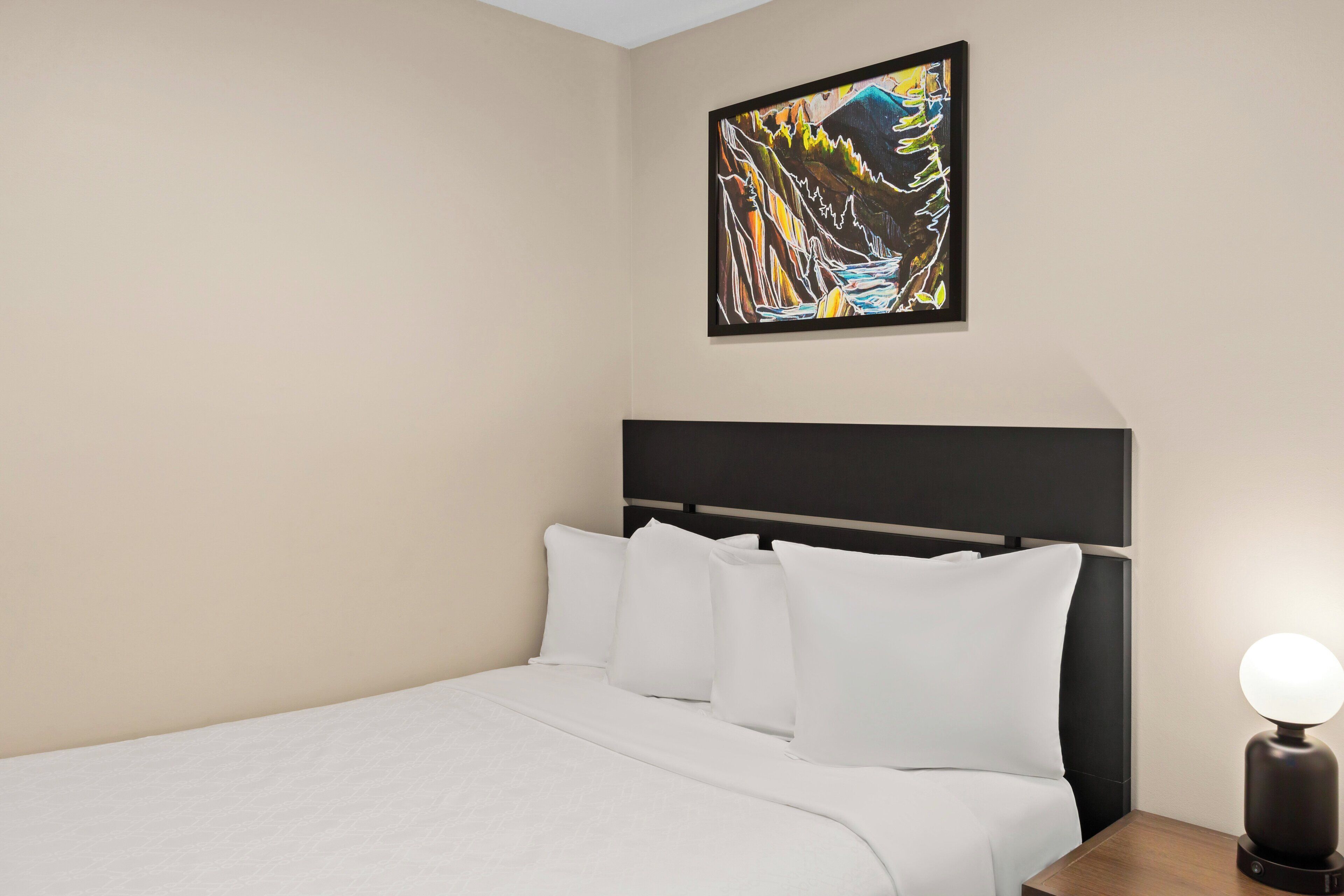 A neatly made bed with white linens and a black headboard, a wall-mounted abstract painting, and a bedside lamp with a white shade.