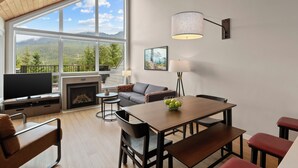 A modern living room with a large window, fireplace, flat-screen TV, sofa, dining table, and red bar stools.