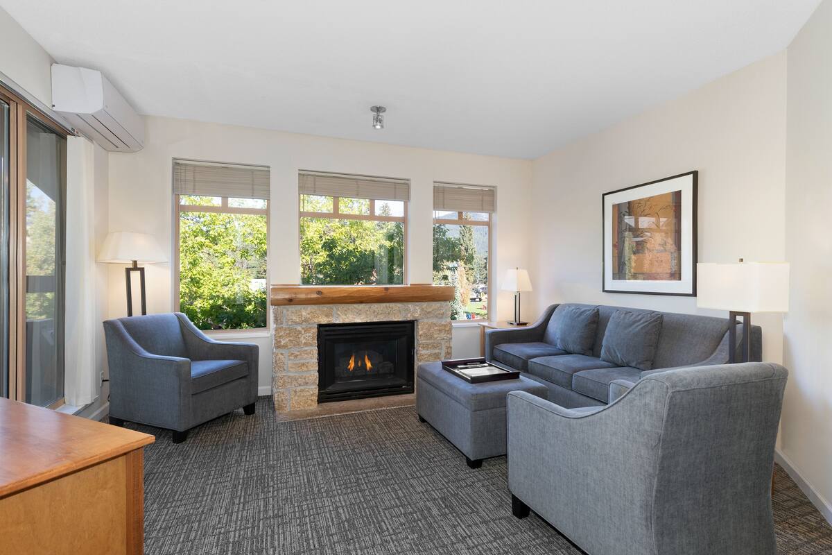 A living room with a fireplace, two grey sofas, a wooden coffee table, and a large window with a view of greenery.