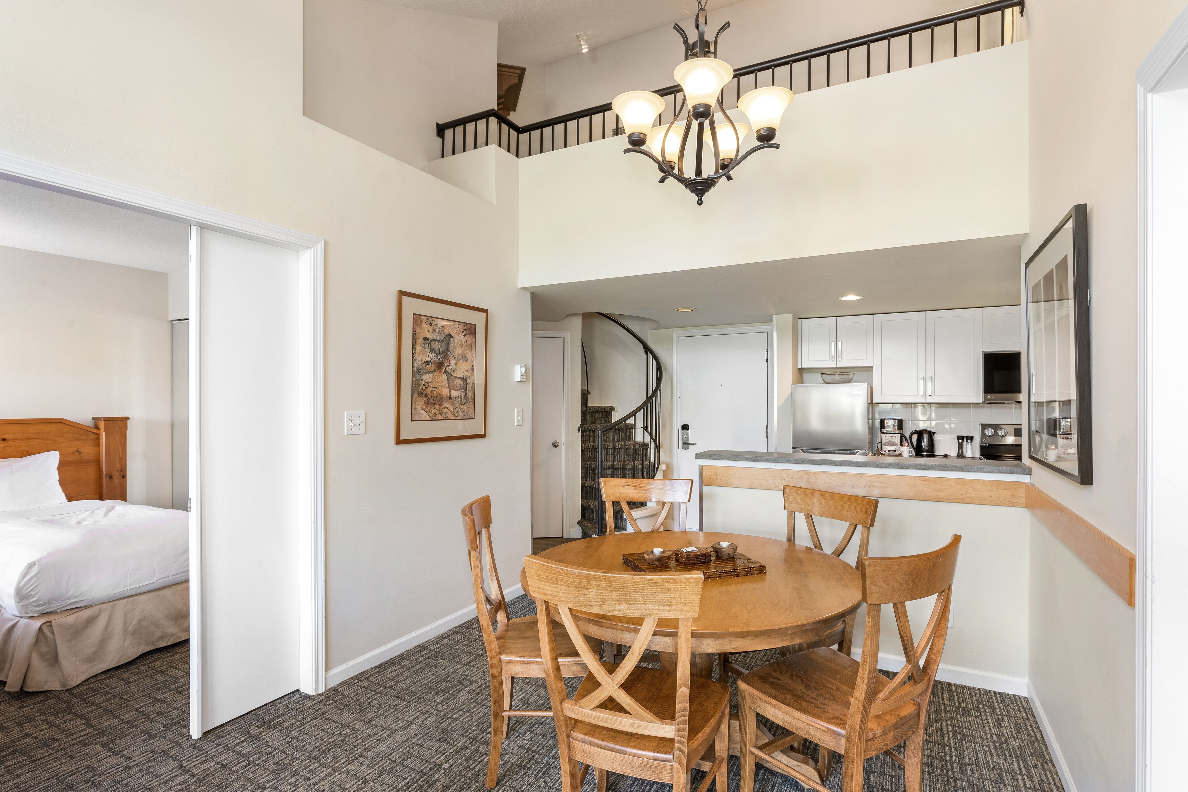 A hotel room with a bed, a dining table with chairs, a kitchen area, and a staircase.