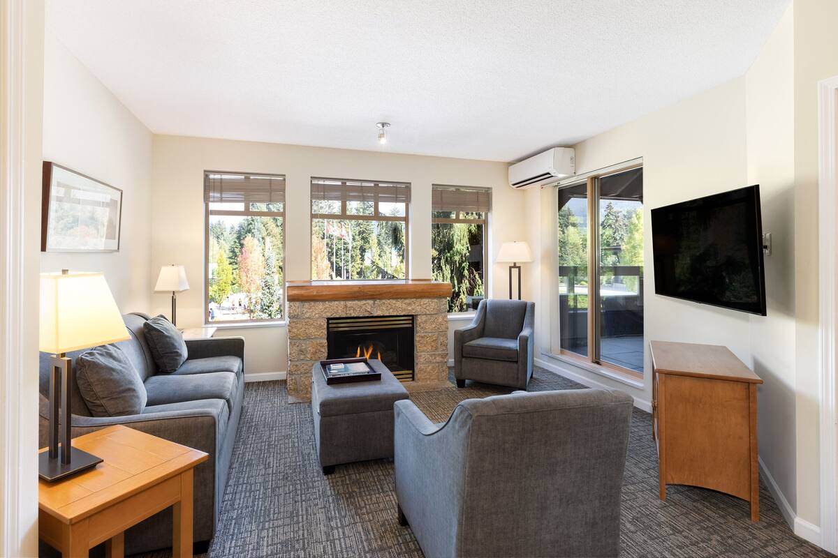 A modern living room with a fireplace, comfortable seating, a flat-screen TV, and large windows offering a view of the outdoors.