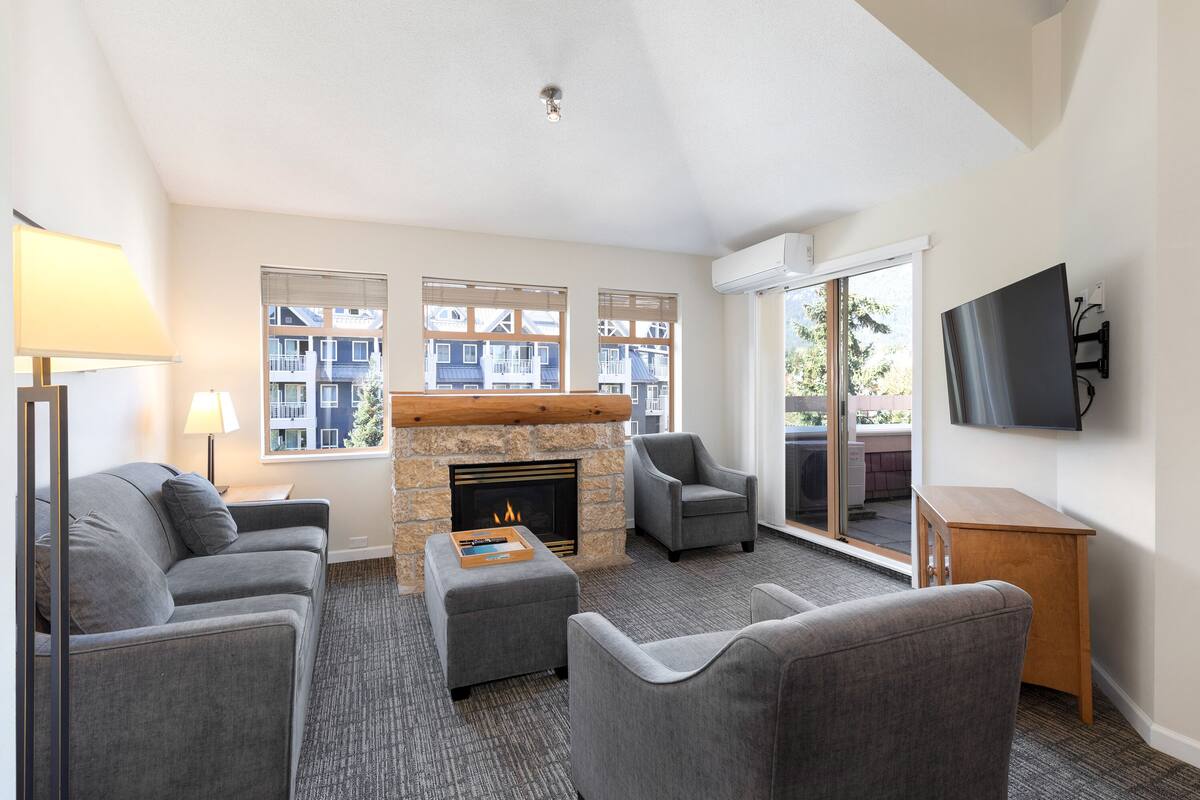 A modern living room with a fireplace, grey sofas, a flat-screen TV, and large windows offering a view of buildings.