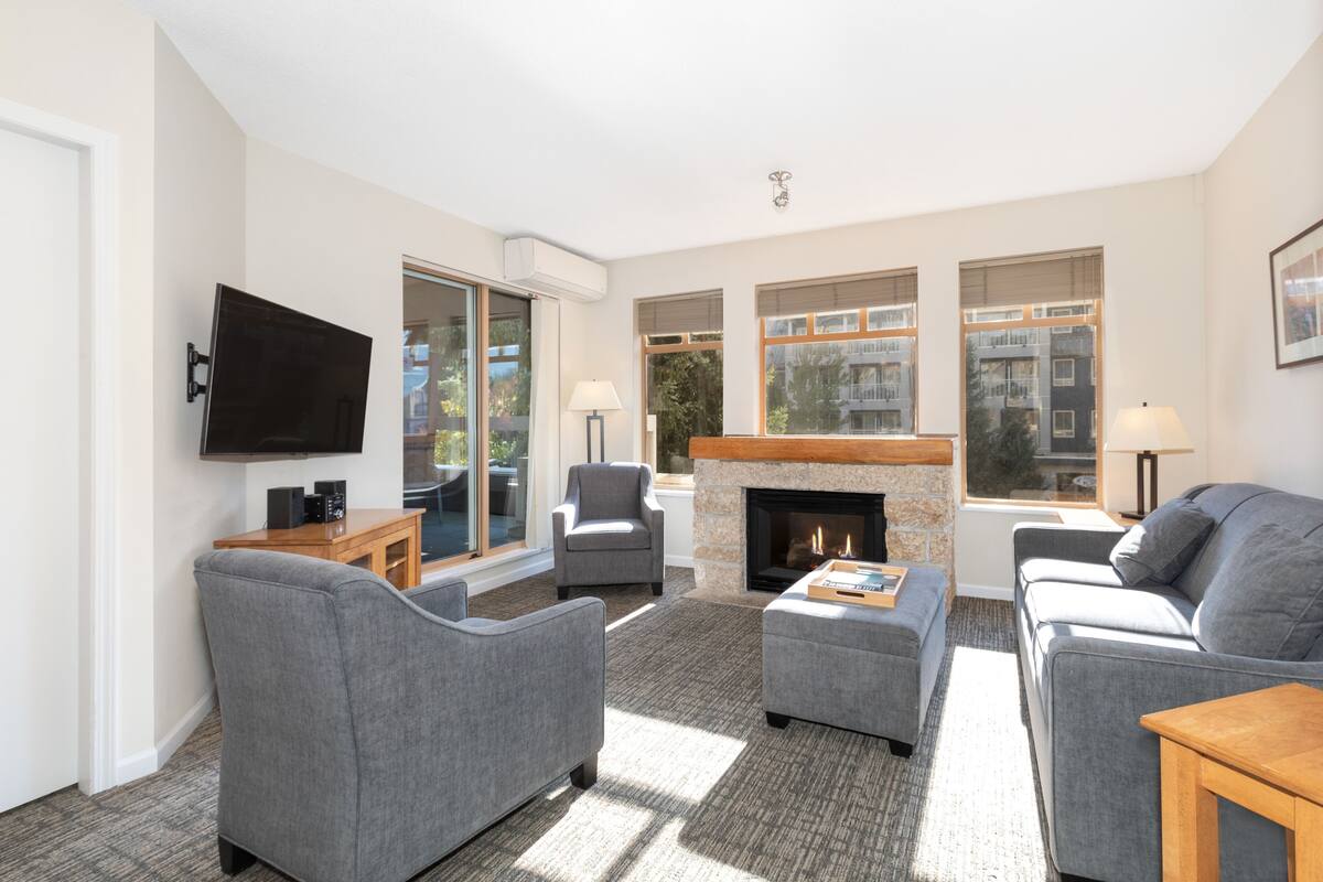 A modern living room with grey sofas, a flat-screen TV, a fireplace, and large windows offering views of the outside.