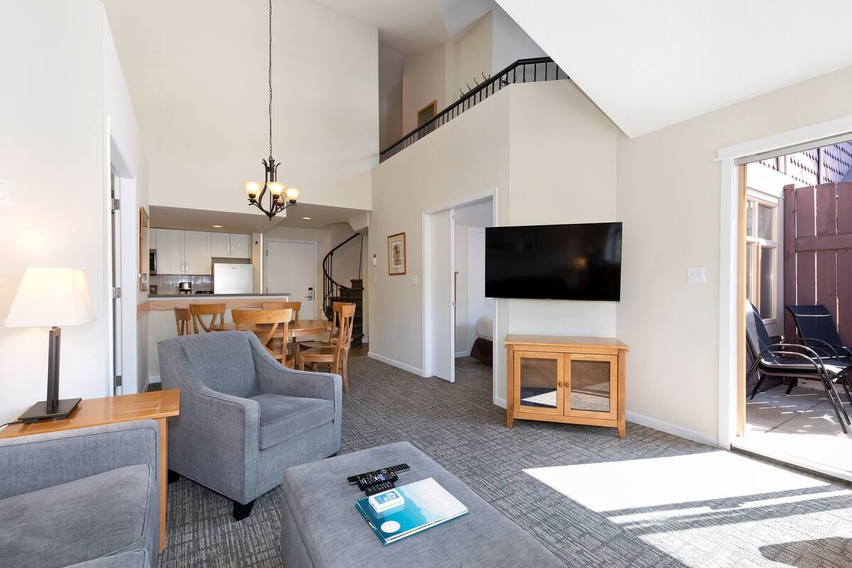 A modern living room with a grey sofa, a wooden coffee table with a book and remote controls, a TV on the wall, and a staircase leading to an upper level.