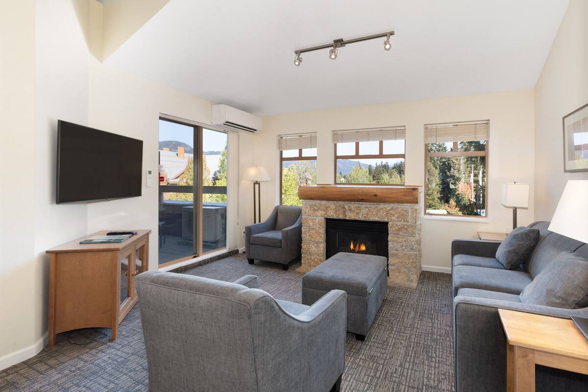 A modern living room with a fireplace, grey sofas, a flat-screen TV, and large windows offering a view of the outdoors.