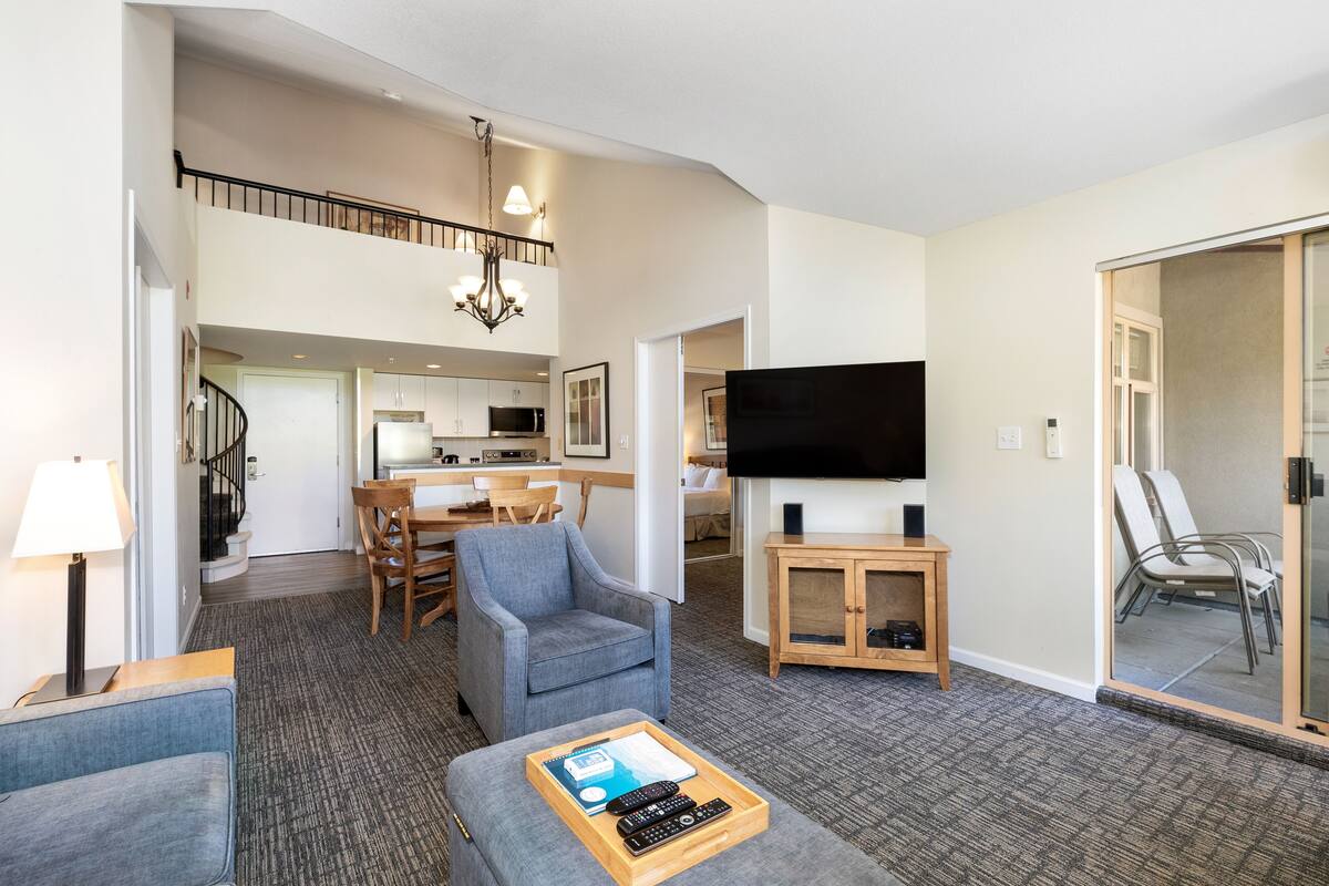 A spacious living area with a grey armchair, a wooden coffee table with remote controls, a flat-screen TV, and a staircase leading to an upper level.