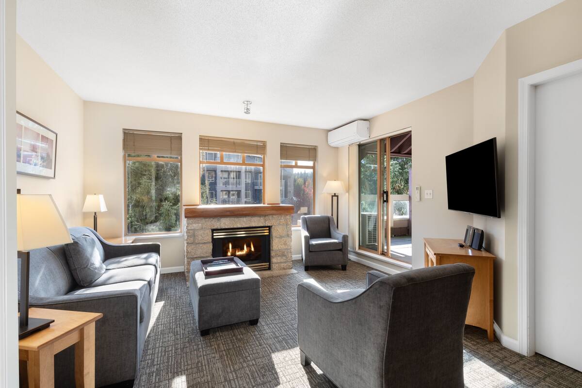 A modern living room with a fireplace, comfortable seating, a flat-screen TV, and large windows offering a view of the outdoors.