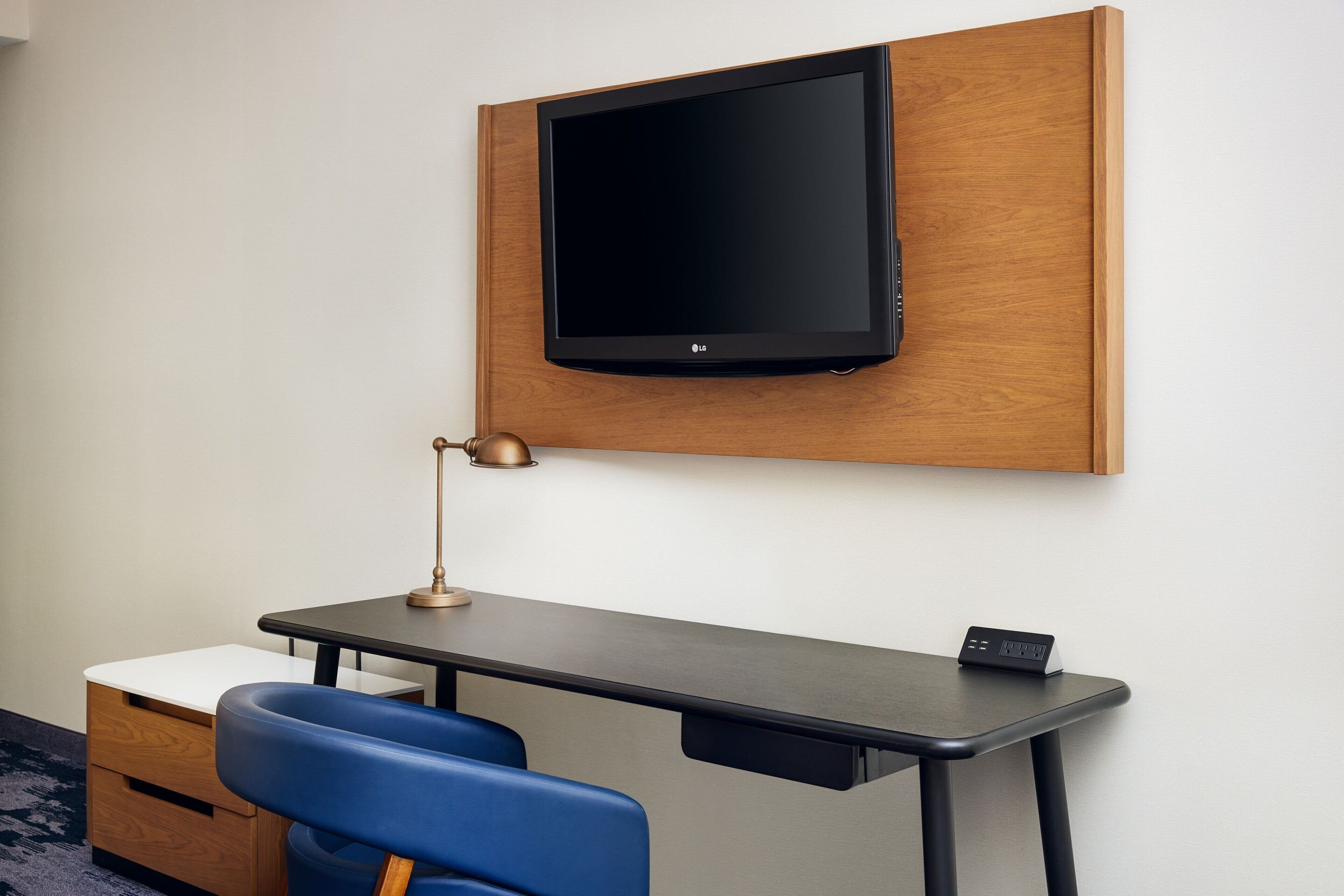 A modern hotel room with a wooden panel wall, a mounted flat-screen TV, a desk with a lamp, a chair, and a small bedside table.