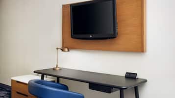 A modern hotel room with a wooden panel wall, a mounted flat-screen TV, a desk with a lamp, a chair, and a small bedside table.