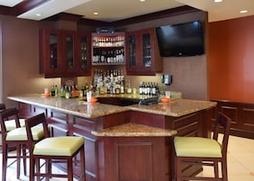 Uma área de bar com bancada de granito, diversas garrafas de bebidas, uma TV de tela plana e banquetas.