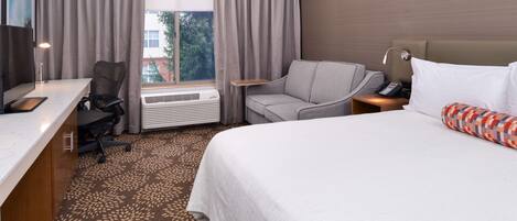 Habitación de hotel con cama, escritorio, silla, sofá y ventana con cortinas.