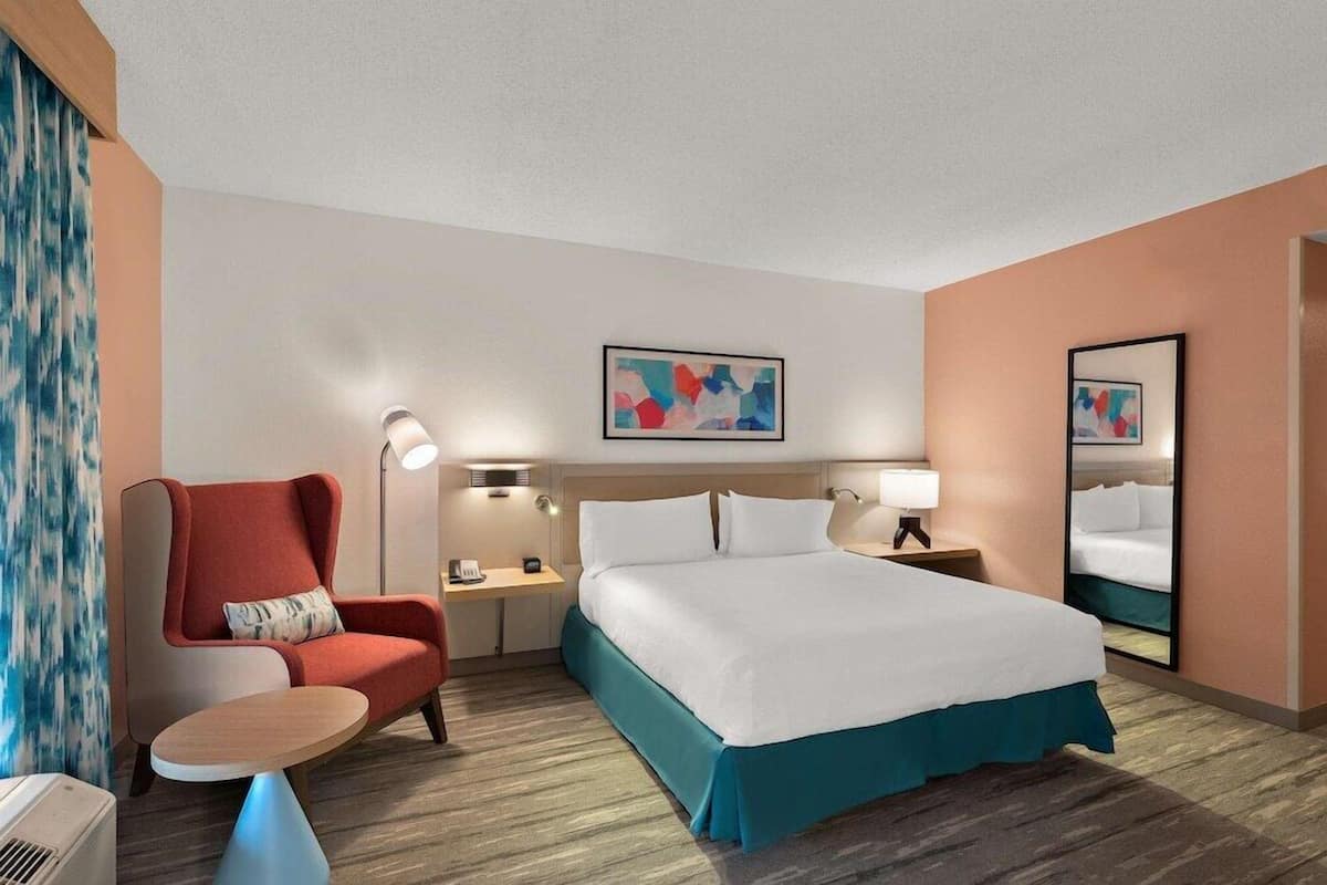 A hotel room with a bed, a chair, a desk, a lamp, and a painting on the wall.