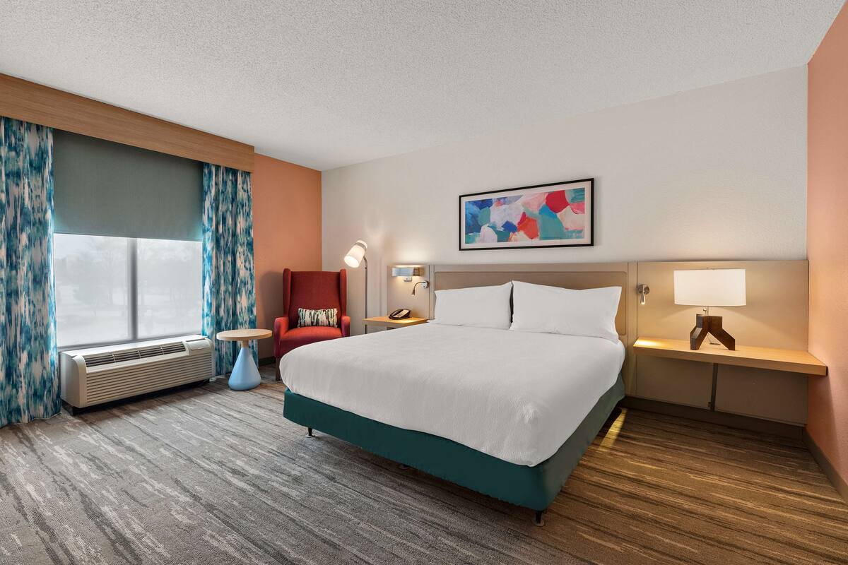 A hotel room with a large bed, a chair, a desk, and a window with curtains.