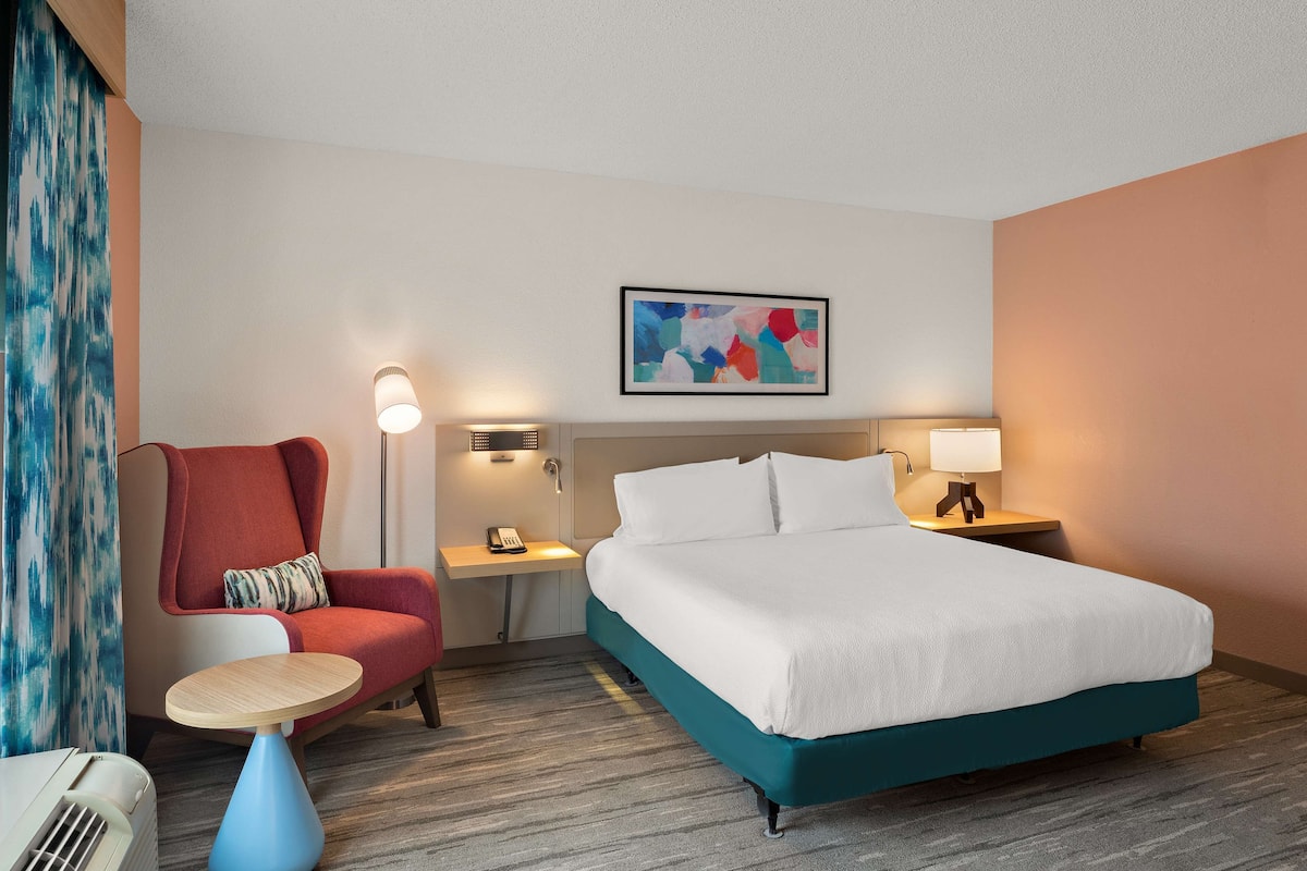 A hotel room with a bed, a chair, a small table, a lamp, and a painting on the wall.
