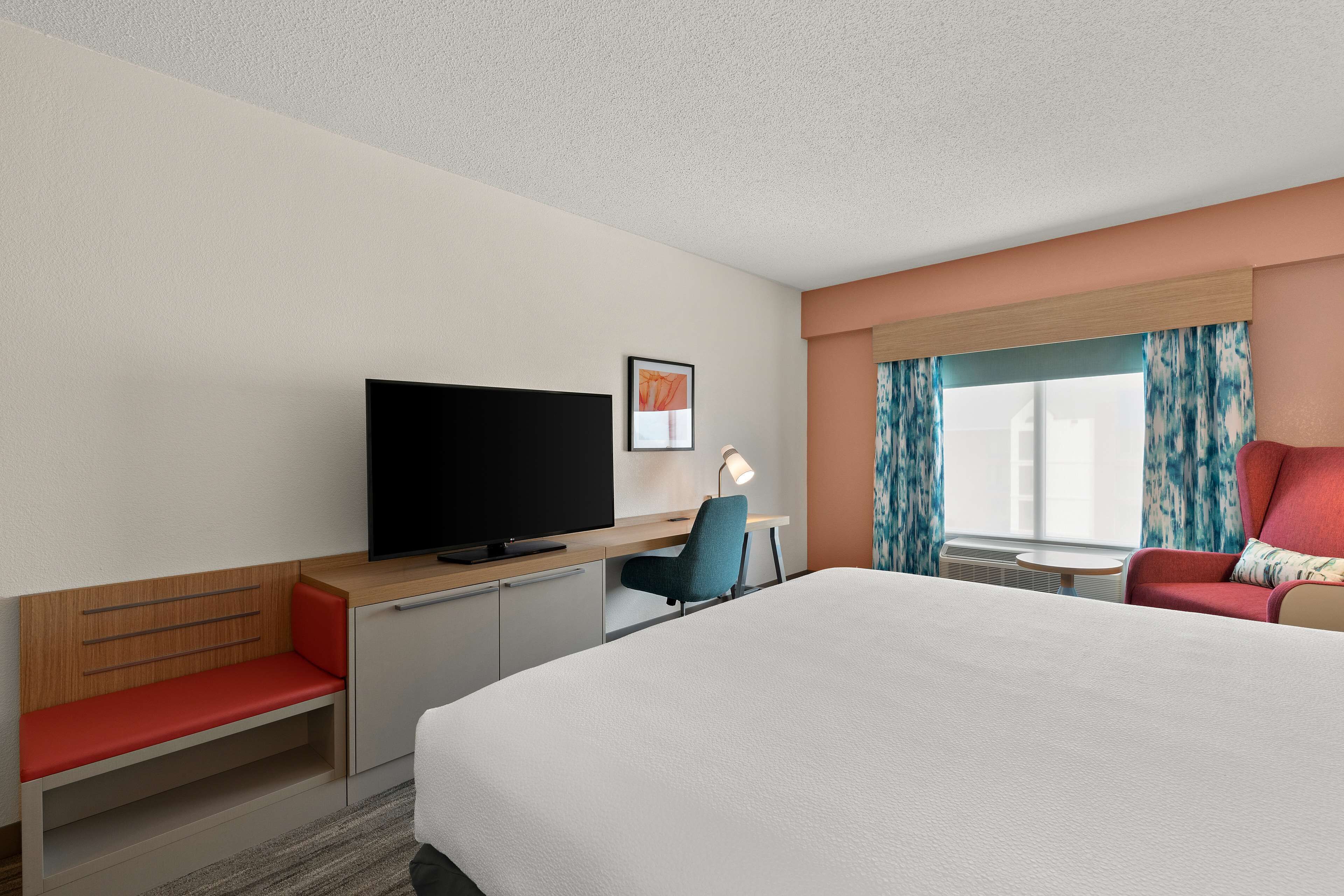 A hotel room with a bed, a desk with a chair, a television, a small table, and a window with curtains.