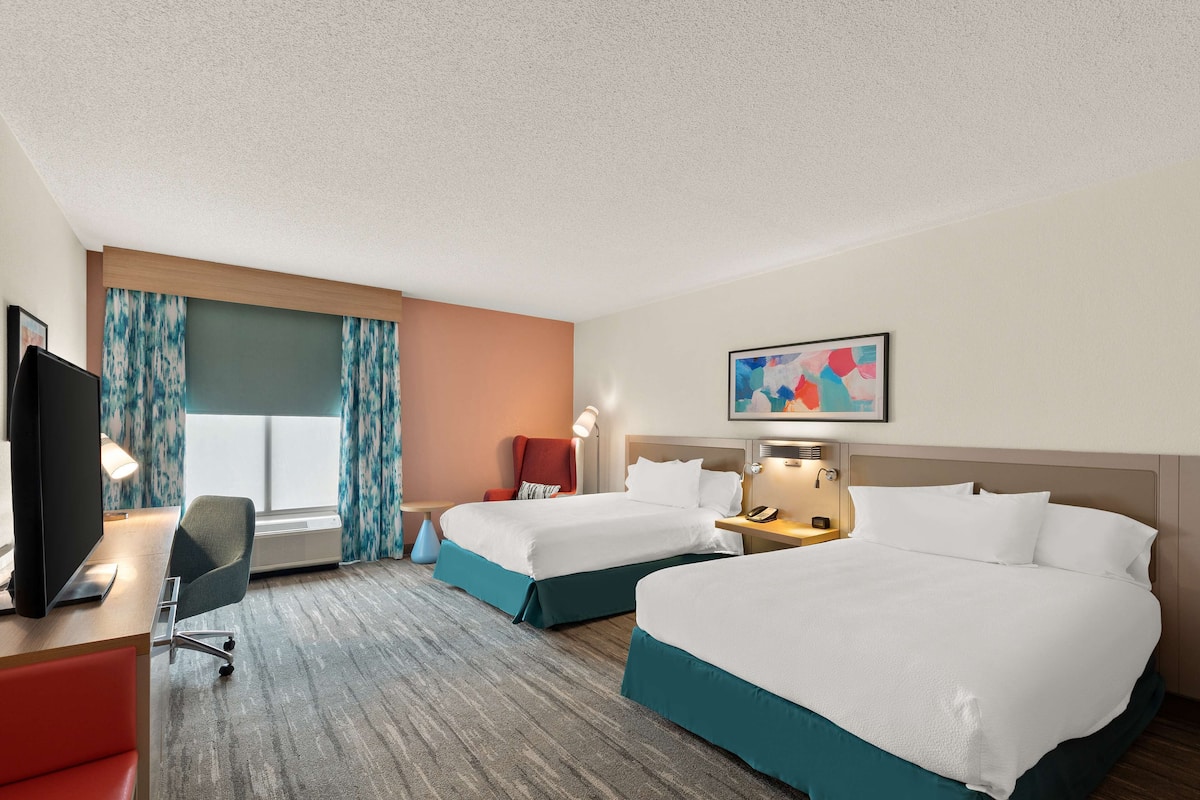 A hotel room with two beds, a desk with a computer, a chair, a TV, and a window with curtains.