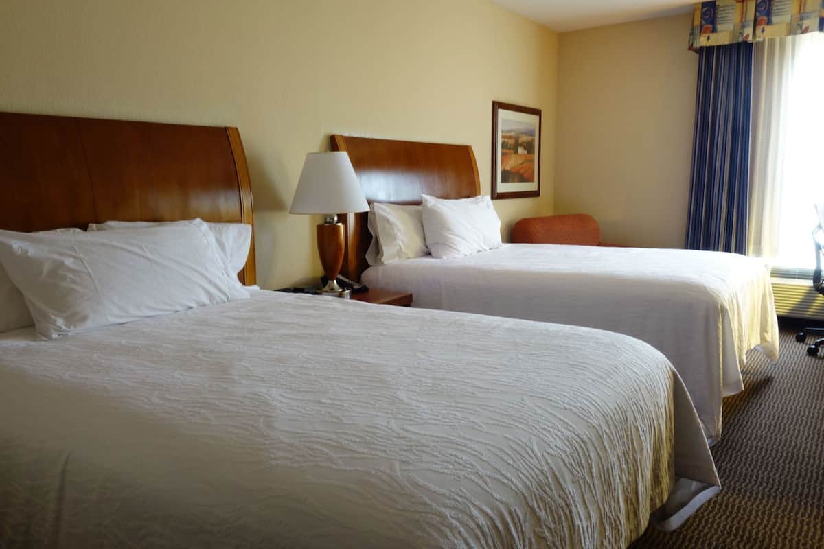 A hotel room with two beds, a nightstand with a lamp, a framed picture on the wall, and a window with curtains.