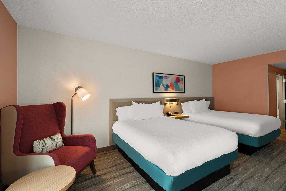 A hotel room with two single beds, a red armchair, a small table, a lamp, and a painting on the wall.