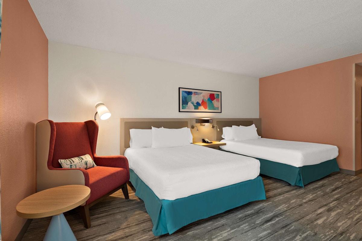 A hotel room with two single beds, a red armchair, a small round table, a nightstand with a lamp, and a painting on the wall.