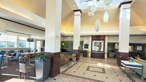 Een ruime lobby met hoge plafonds, grote ramen en een centraal zitgedeelte met tafels en stoelen.