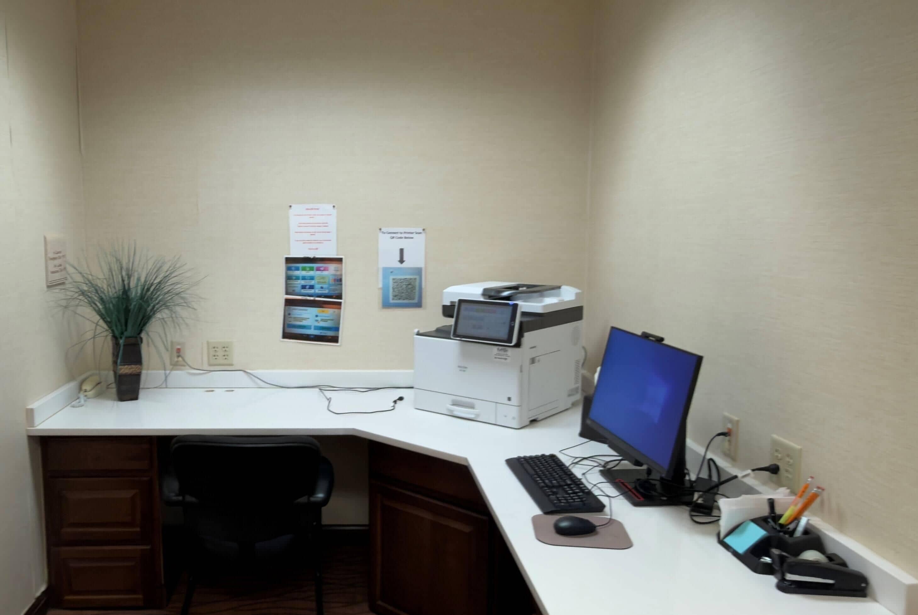 A corner office with a desk, computer, printer, and a potted plant.