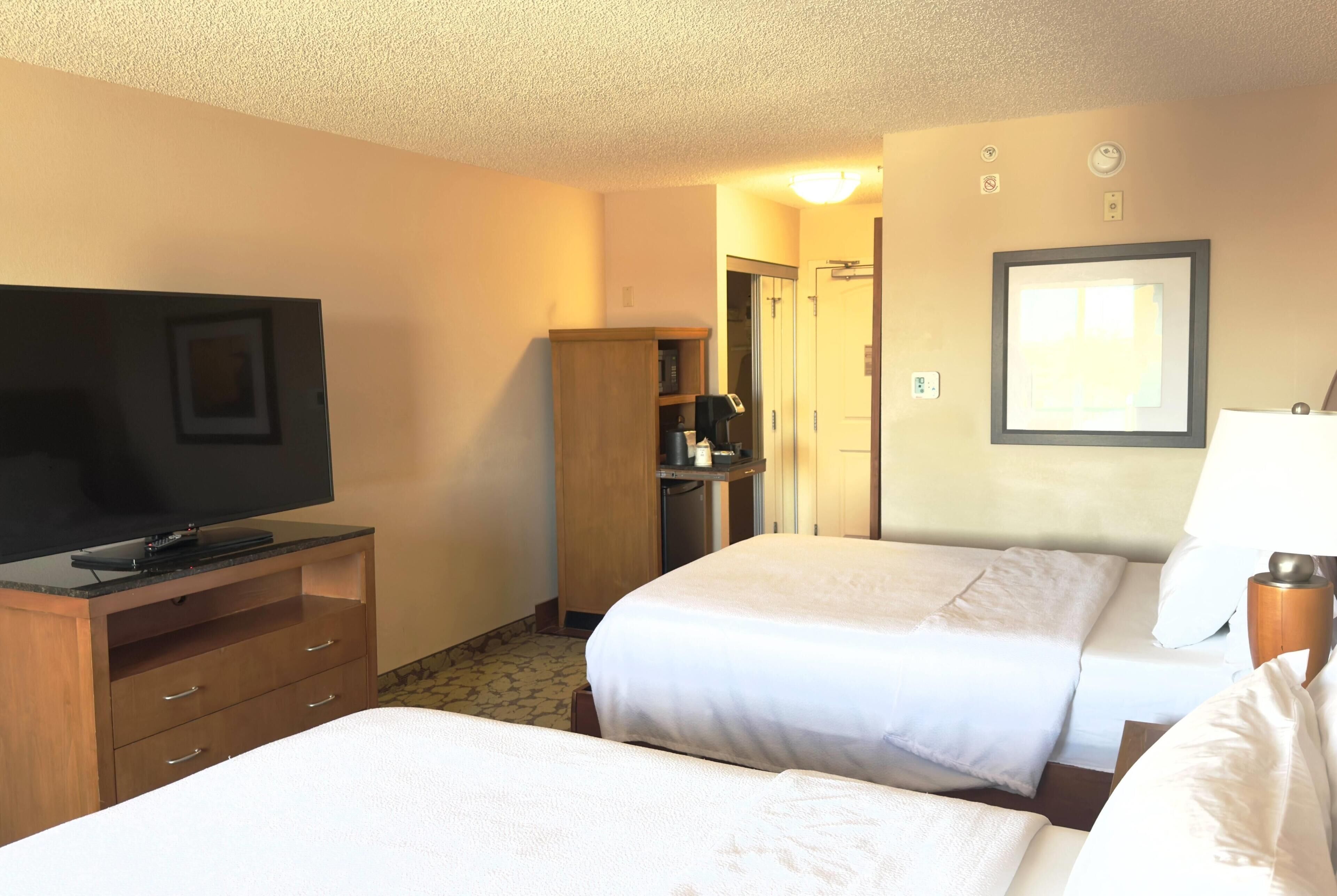 A hotel room with a television on a wooden cabinet, two beds with white linens, a wooden wardrobe, and a framed picture on the wall.